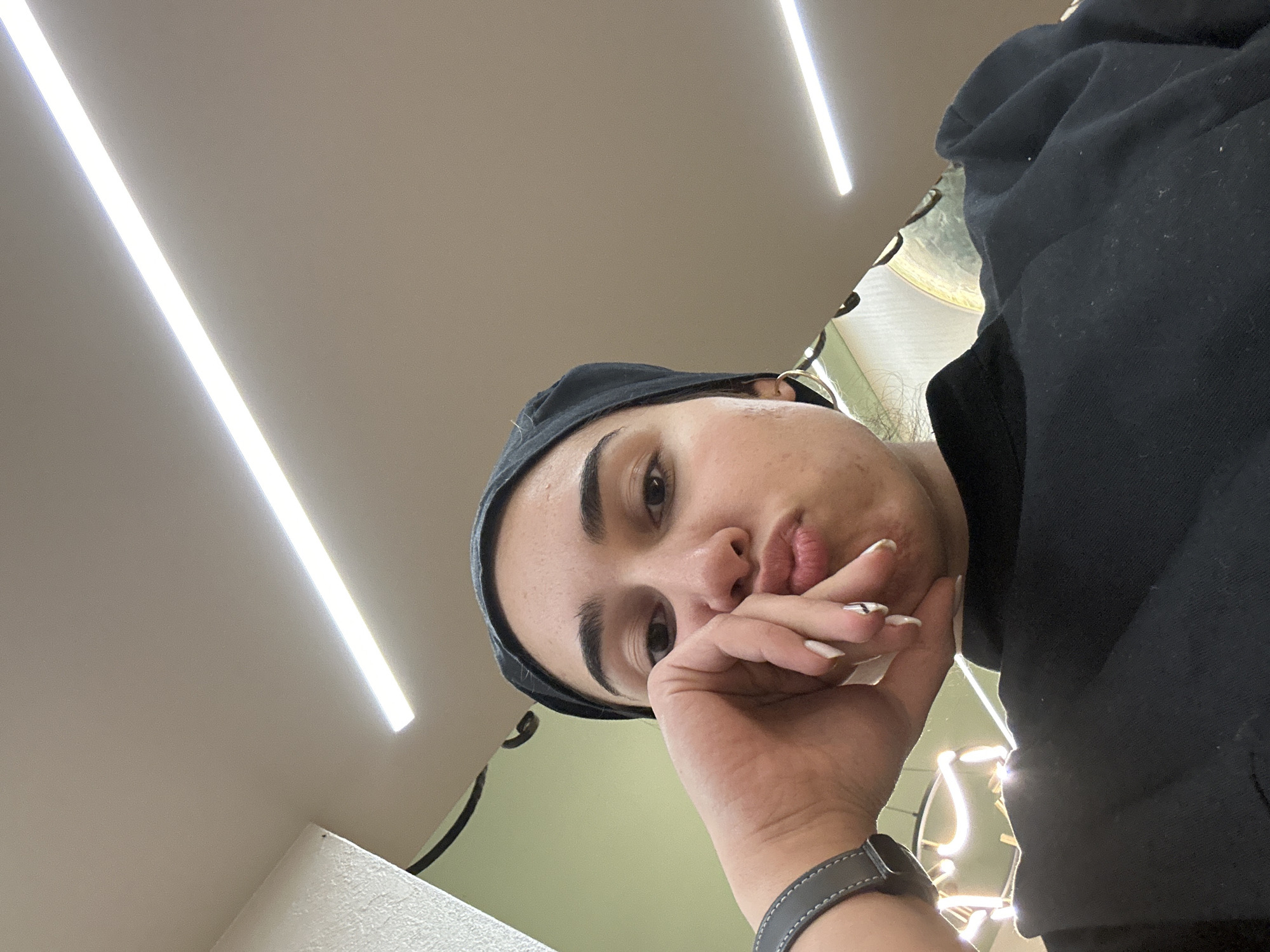 A woman with dark hair is wearing a black cap and a black hoodie. She has light skin and is wearing a grey watch. Her nails are painted white with a black design. The background includes a white ceiling with two bright linear lights, a light green wall, and what appears to be some kind of decorative lighting. The location is likely indoors. It is not possible to confirm whether the location is in Burgas, Bulgaria.