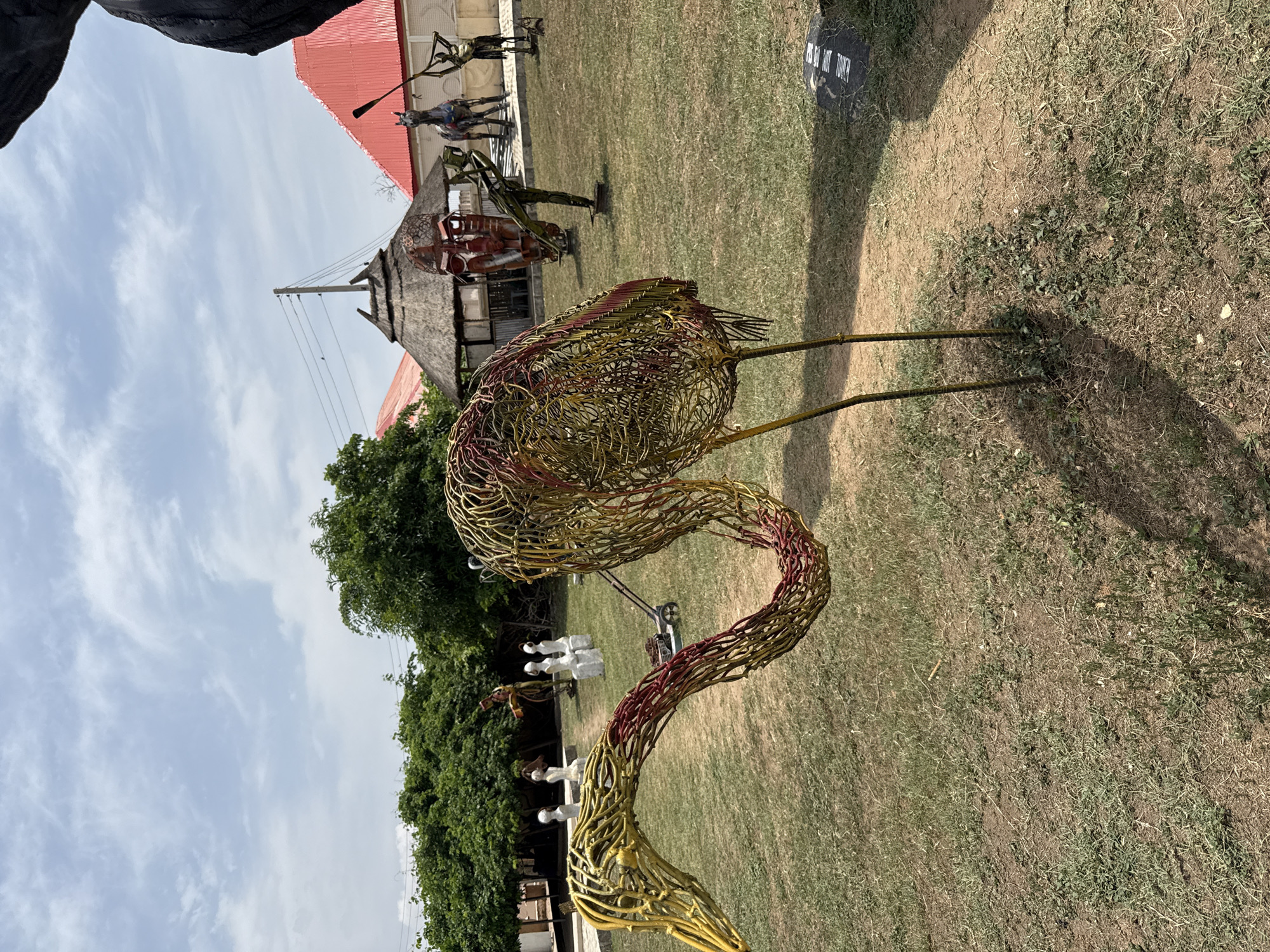 The image displays an outdoor sculpture garden or art park under bright daylight with a partly cloudy sky. In the foreground, a large, intricate sculpture of a bird, resembling a flamingo or crane, stands prominently. It is crafted from woven wire, primarily yellow and reddish-brown, forming its body, long curving neck, head, and thin legs.
In the middle ground, several other striking sculptures are visible. These appear to be made from recycled metal or wire and depict human-like figures in various dynamic poses; one is on horseback, another holds a spear-like object, and others stand erect. Behind these sculptures, there are buildings, including one with a traditional thatched roof and another with a red corrugated metal roof. A utility pole with overhead wires is also visible. Further in the mid-ground, near the bird's neck, several abstract white sculptures (possibly stools or small figures) are arranged.
The ground is covered with sparse, dry grass and dirt, with some patches of greener foliage. Lush green trees and bushes frame the background and the left side of the scene. Strong shadows cast by the sculptures and buildings indicate a high sun. A black sign on the ground features partially visible white text that reads "IS DO NOT TOUCH," likely indicating a display instruction. The overall scene suggests a public art exhibition space in a sunny, possibly semi-arid environment, consistent with Abuja, Nigeria.