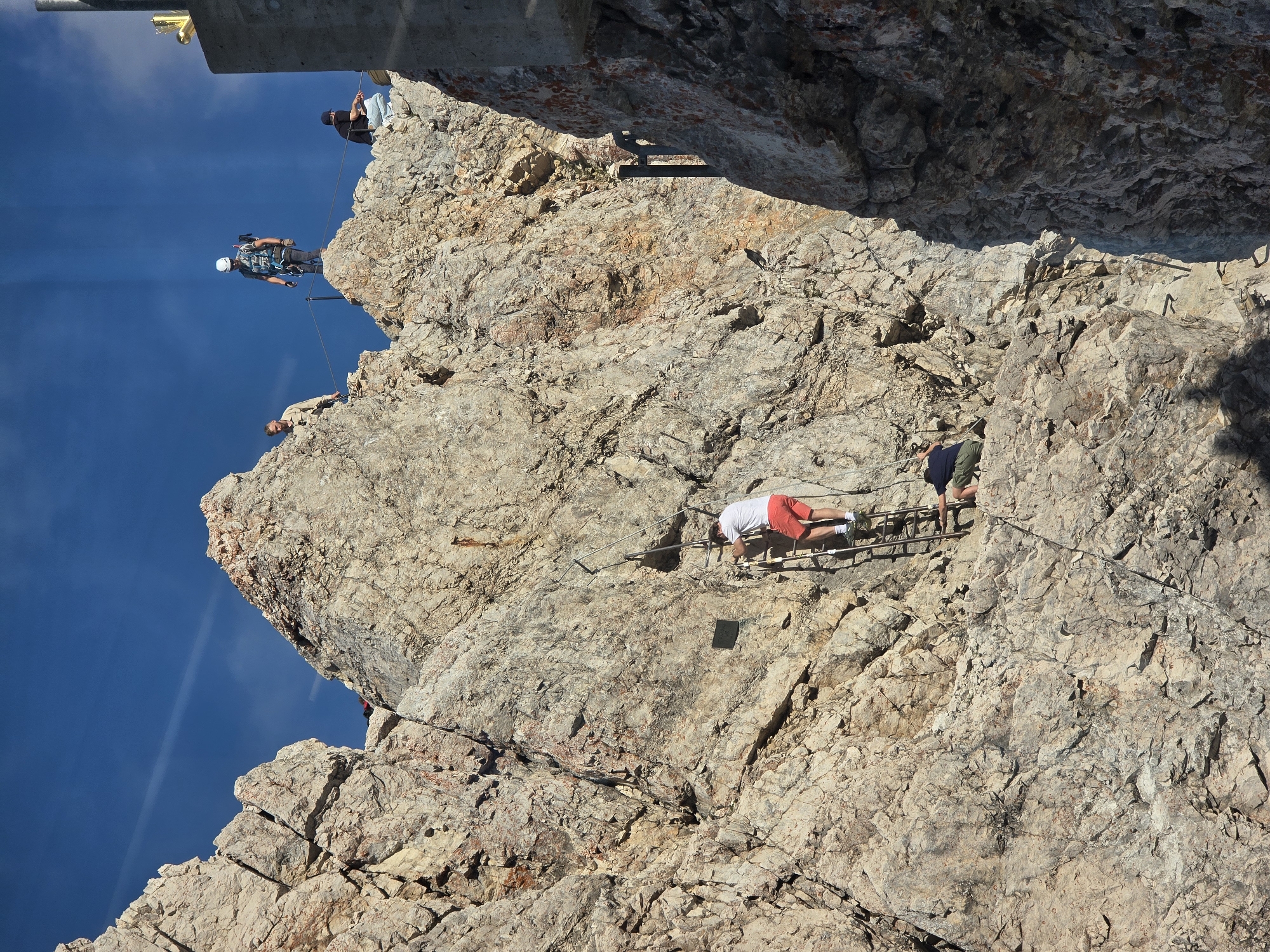 The image captures a group of people engaged in climbing a rocky mountain in Grainau, Germany. The scene is set against a clear blue sky, with the sun casting harsh shadows on the rugged terrain. In the foreground, a man in red shorts and a white t-shirt is ascending a metal ladder bolted to the rock face. Below him, another person in a dark shirt and green pants is climbing a less steep section of the mountain. Higher up on the ridge, several other individuals are visible. One person, wearing a helmet and safety harness, stands on a rocky outcrop, holding onto a rope. Another person in a black t-shirt and blue jeans is seated on the rocks, and a third person in a tan shirt is seen on a different rocky ledge. The mountain itself is characterized by its rough, textured surface with various shades of grey and brown rock, interspersed with patches of reddish-orange mineral deposits. The presence of a metal ladder and safety ropes suggests this is a designated climbing route or via ferrata.