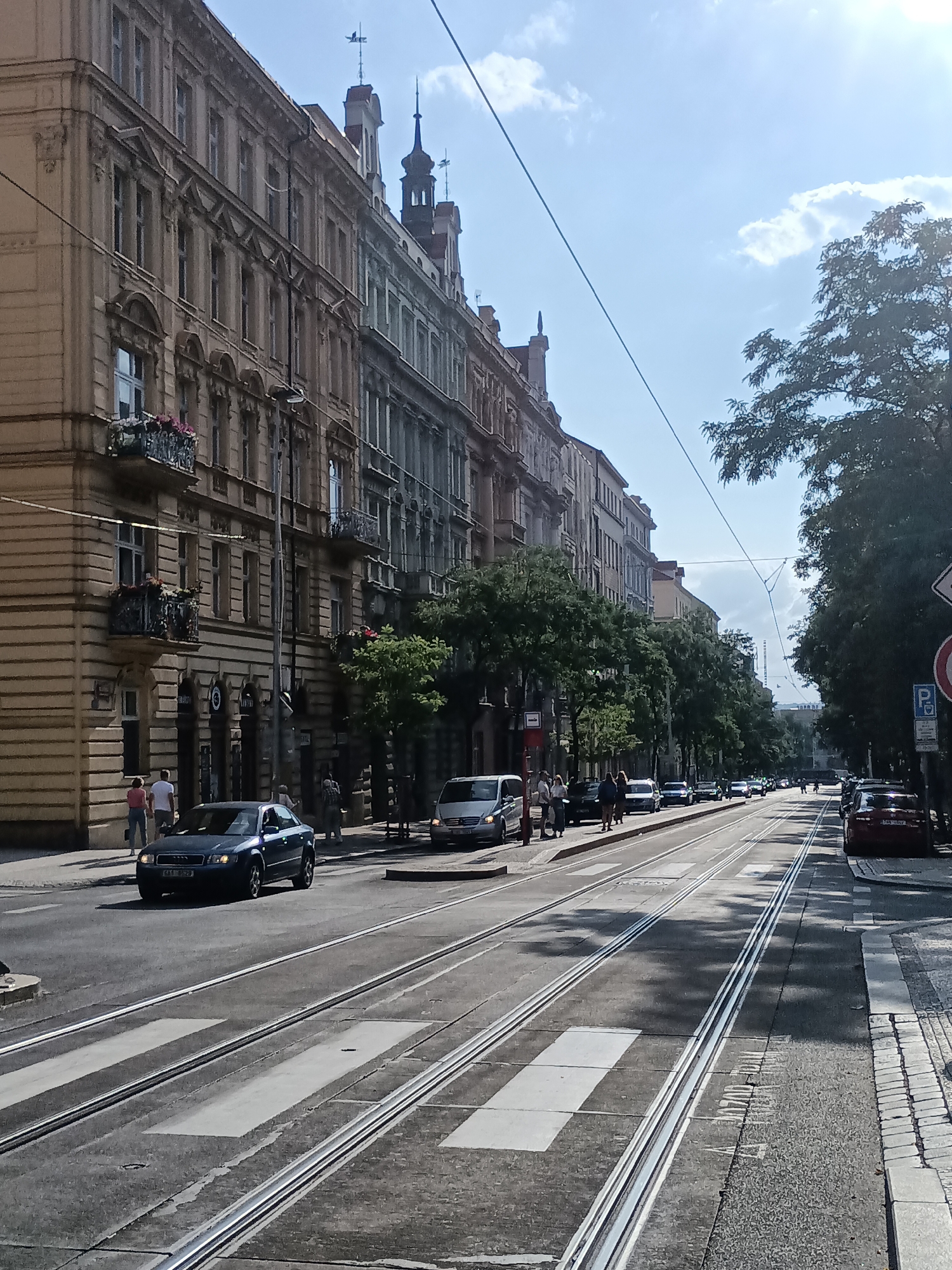 The image captures a street scene in Prague, Czechia, on a sunny day with a clear sky and some scattered clouds. The perspective is from street level, looking down a wide avenue lined with ornate, multi-story buildings on the left. These buildings feature classical architectural elements such as decorative facades, balconies with flower boxes, and arched windows.
The street itself is paved with asphalt and features tram tracks running down the center. Pedestrian crosswalks are visible across the tram tracks. Several cars are present on the road, including a dark sedan in the foreground, a silver minivan to its right, and other vehicles further down the avenue.
On the left side of the street, near the buildings, pedestrians are walking along the sidewalk. Trees with lush green foliage are interspersed along the street, casting shadows on the road. On the right side of the image, more trees and a sidewalk with cobblestone edging are visible, along with traffic signs, including a "P" for parking and a "no parking" symbol. The overall impression is of a busy urban environment in a historic European city.