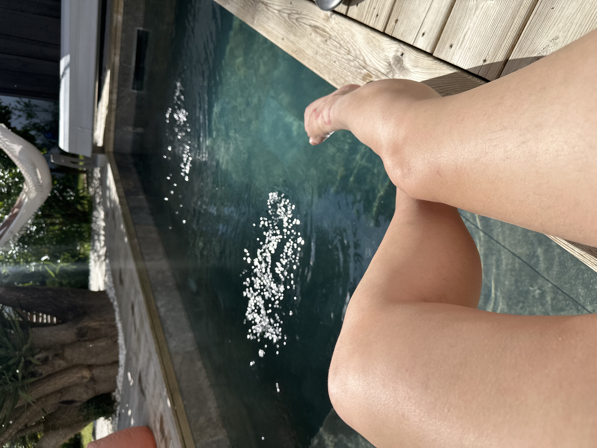 The image shows a pair of human legs dangling over the edge of a pool. The pool's edge is made of light-colored wooden planks. The water in the pool appears dark, with light reflecting off its surface, creating bright spots. In the background, there are plants and a glimpse of a white hammock. Based on the location information, the scene is in La Couarde-sur-Mer, France.