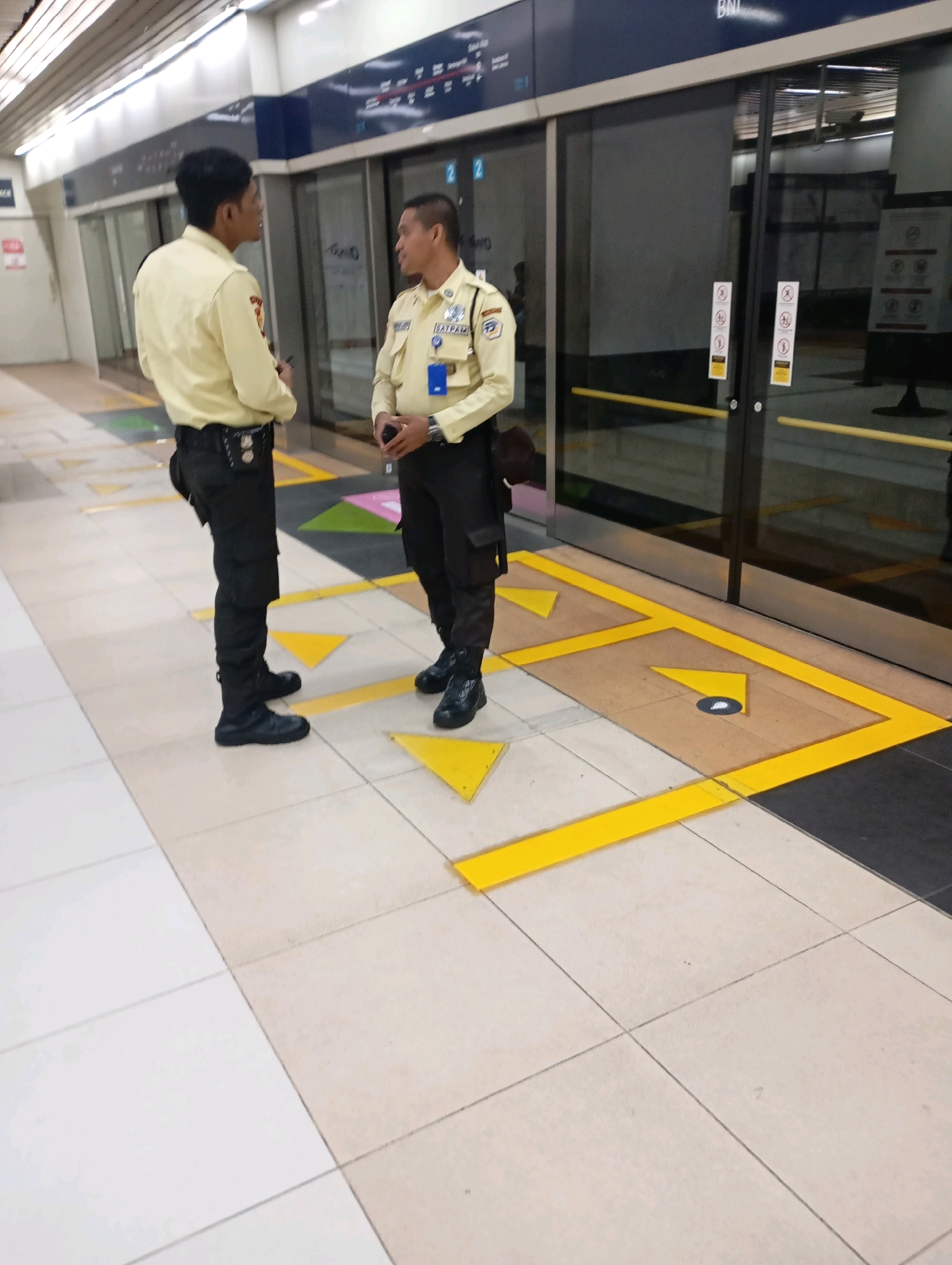 Two male security guards in yellow shirts and black pants stand on a train platform in Central Jakarta, Indonesia. The guard on the left, facing away from the viewer, is speaking to the guard on the right, who is looking at him and holding a dark object in his hands. Both guards are wearing black boots. They are standing on a tiled platform with yellow directional arrows painted on the floor. Behind them are glass doors leading into a train, with advertisements visible inside. The train station appears to be modern and well-lit.