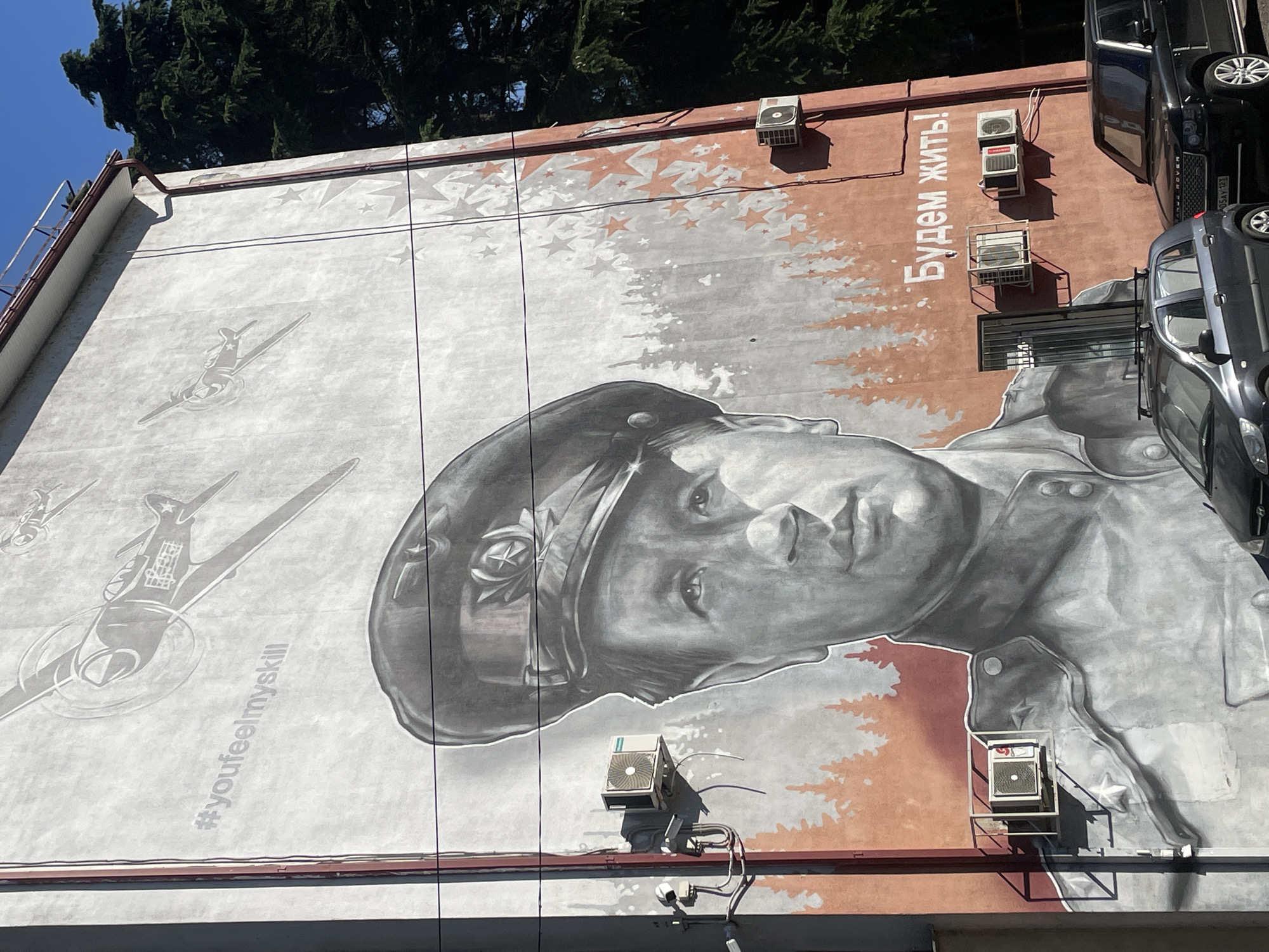 The image shows a mural on the side of a building in Sochi, Russia. The mural depicts a grayscale portrait of a man in a military uniform and cap, with stylized trees and stars in the background. Above the portrait, there are images of airplanes. Text in English reads "#youfeelmyskill", and text in Russian reads "Будем жить!" (We will live!). Several air conditioning units are mounted on the wall of the building, along with some electrical wiring and what appears to be a security camera. Two cars are parked in front of the building, a dark gray hatchback and a black SUV. Evergreen trees are visible to the right of the building. The sky is clear and blue.