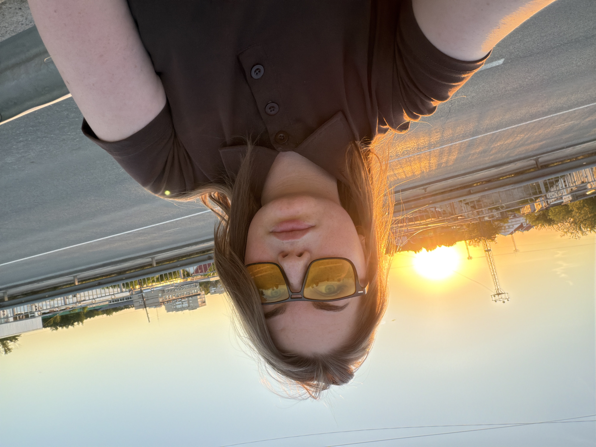 A woman is taking a selfie on a bridge in Chaikovsky, Russia, at sunset. She is wearing a brown collared shirt and yellow-tinted glasses. The sun is directly behind her, creating a halo effect around her hair. The cityscape of Chaikovsky can be seen in the background, including buildings and a weather station. The bridge appears to be over a roadway.