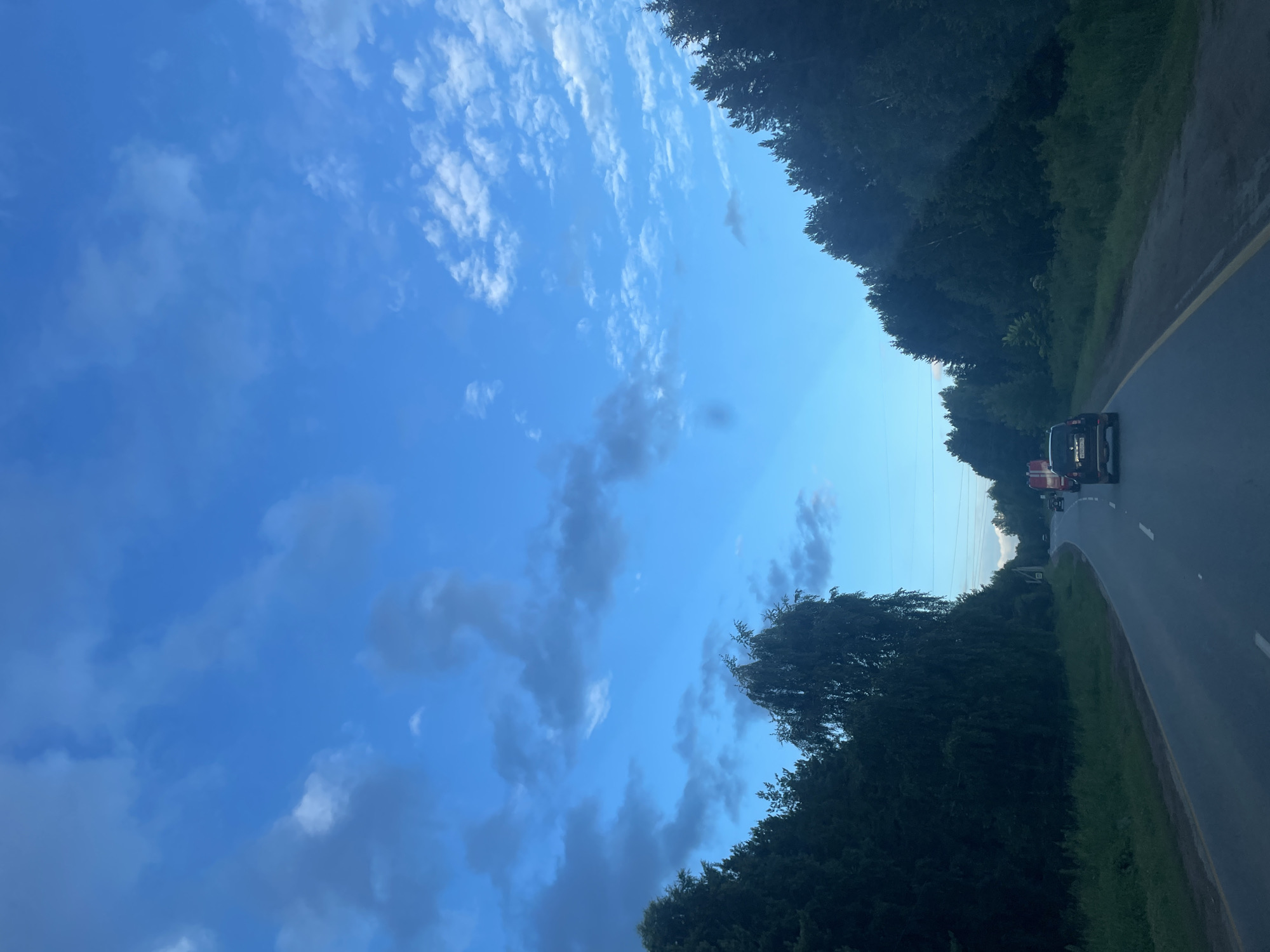 The image depicts a roadside scene in the vicinity of Kamkoye, Russia. The perspective is from a moving vehicle looking towards the right side of the road. The time of day appears to be late afternoon or early evening, judging by the soft light and the presence of scattered clouds against a bright blue sky. The weather seems pleasant, with clear skies and a gentle breeze suggested by the wispy clouds.
The primary subjects are a dark-colored SUV and a red vehicle, possibly a truck or van, driving ahead on a paved road. The road is bordered by a yellow line on the right and a grassy shoulder. Dense, dark green trees line the left side of the road, creating a natural barrier. In the background, more trees and vegetation are visible, suggesting a rural or semi-rural setting. A faint hint of a power line can be seen stretching across the horizon.
No people are clearly visible, but the presence of vehicles implies human activity. The overall mood is one of travel and transition, with the open road and the natural landscape contributing to a sense of journey. The vibrant blue sky and the lush greenery evoke a feeling of natural beauty and tranquility. There is no discernible text in the image.