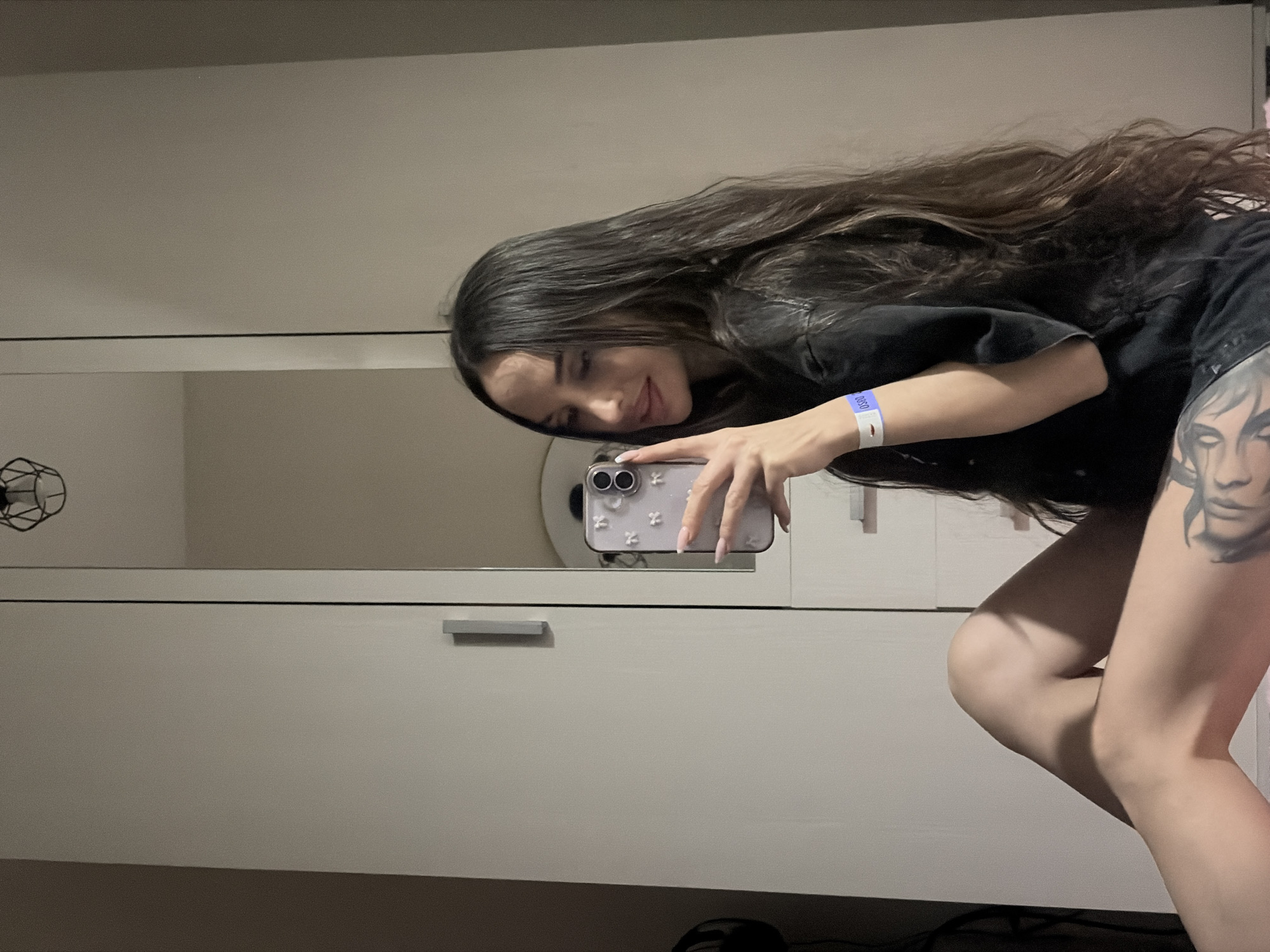 A young woman with long, dark hair is taking a selfie in front of a light-colored wardrobe. She is wearing a dark-colored shirt and shorts, and has a tattoo on her thigh. She is holding a smartphone with a lilac case. The environment suggests an interior space. There is no verifiable information to suggest that this image was taken in Saint Petersburg, Russia.