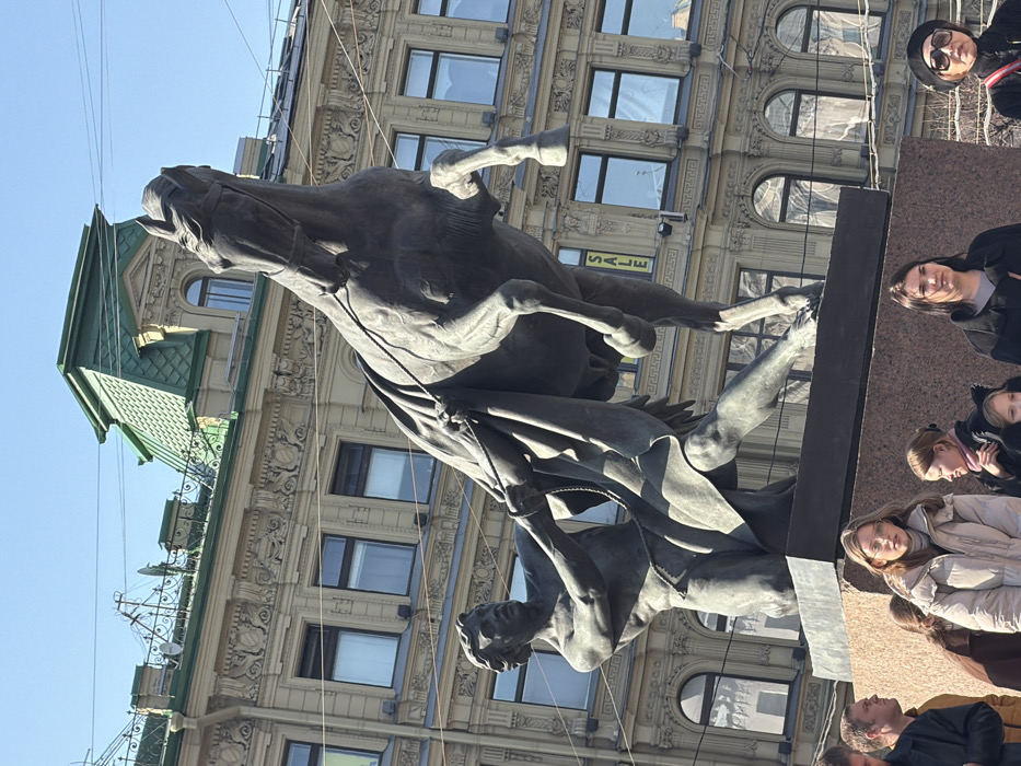 The image presents a rotated view of a grand equestrian statue set against the backdrop of an ornate city building on a clear, sunny day. The camera appears to be pointed upwards, capturing the statue from a low angle.
The central subject is a large, dark-colored, likely bronze, statue depicting a dynamic and dramatic scene. It features a powerful, rearing horse with its front legs lifted high off the ground, its mouth open in an apparent neigh or struggle. Below the horse, a muscular, classically sculpted man is depicted grappling with the animal, holding onto reins or ropes. The statue conveys a sense of strength, tension, and movement. It rests upon a dark, rectangular pedestal, partially visible on the right side of the frame.
Behind the statue, a multi-story, light-colored building with elaborate architectural details rises into the sky. The building features numerous windows, some rectangular and others with arched tops on the lower visible floors. Decorative elements such as pilasters, cornices, and detailed friezes adorn the facade. On the far left of the building, a green, pitched or mansard-style roof with dormer windows and some metalwork details is visible. Several thin wires, possibly electrical or tram lines, crisscross the clear blue sky and pass in front of the building.
A clear, bright blue sky fills the upper left portion of the image, indicating sunny weather.
To the right of the statue and its pedestal, several people are visible, some looking towards the statue. In the top right corner, a person wearing sunglasses and a dark jacket is partially visible. Below them, a woman with dark hair in a black top is looking in the direction of the statue. Further down and slightly more in the foreground, a group of individuals, including children and adults dressed in winter coats and scarves, are present, seemingly observing the monument or walking by. Their expressions appear neutral and observant.
The only visible text is the word "SALE" in yellow letters on a dark sign, positioned vertically and reversed on the building behind the horse's torso. The setting is clearly an urban environment, likely a public square or a wide street, suggested by the scale of the statue and the surrounding architecture.