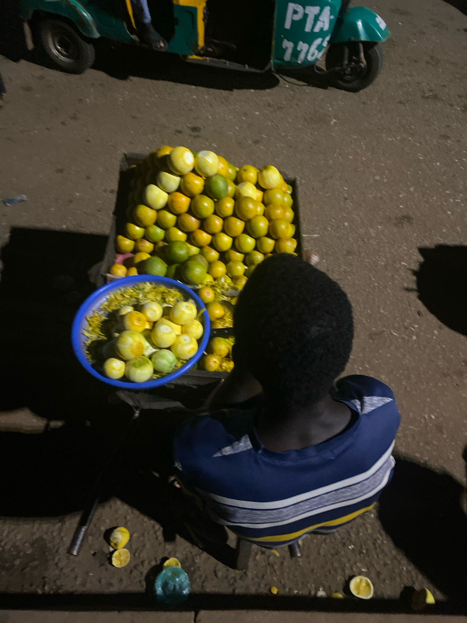 An overhead, medium shot captures an individual from behind, seated in front of a cart overflowing with citrus fruits. The person wears a blue and white striped shirt with yellow accents. In front of them is a blue bowl filled with what appears to be peeled oranges and yellow flower petals. To the left of the bowl, the cart holds a dense arrangement of oranges, some green and some yellow. The cart itself is an old, weathered wooden structure on wheels. Behind the cart, a three-wheeled vehicle, a "Keke Napep," is partially visible, painted green and yellow with "PTA 776" written on its side. The ground is paved with rough, dark asphalt. Scattered on the ground near the cart are a few discarded pieces of citrus fruit, one of which has been sliced in half. The scene appears to be a street-side market, likely in the city of Bukuru, Nigeria, given the presence of the Keke Napep and the general atmosphere. The lighting suggests it is either early morning or late evening.