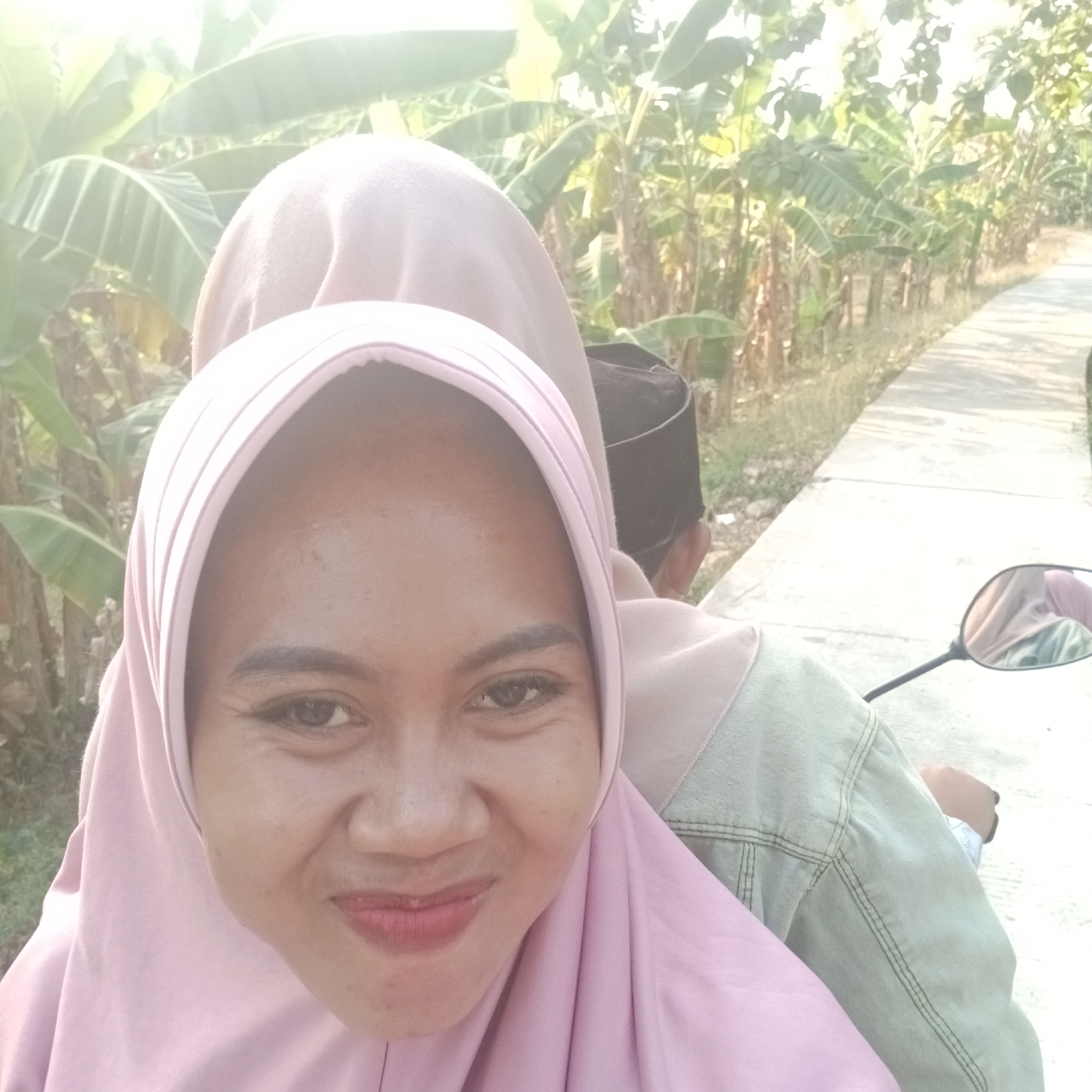 The image shows a close-up selfie of a young woman wearing a pink hijab and a light-colored denim jacket. She is on the back of a motorcycle, with another person wearing a black skullcap visible behind her. The woman is smiling, and her lips are painted with pink lipstick. Her right eye is visible, and she has defined eyebrows. The background features lush green banana trees and a paved road, suggesting a rural or semi-rural setting. The sun is bright, casting shadows and creating a slightly overexposed effect, particularly in the upper part of the image. The reflection in the motorcycle's side mirror shows a glimpse of the people on the motorcycle and the surrounding foliage. The scene is captured in Tegowanu Kulon, Indonesia, as indicated by the provided context.