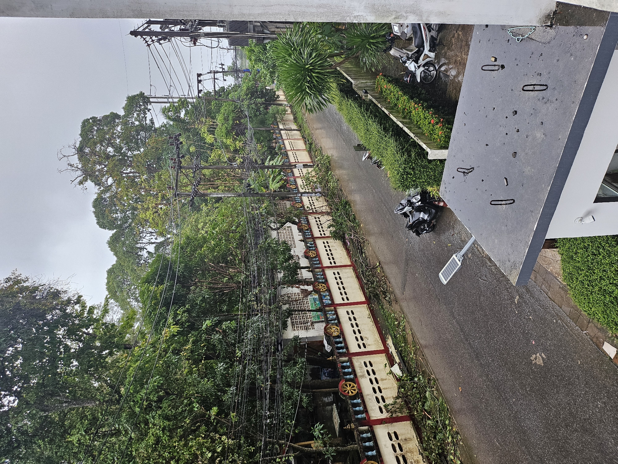 The image shows a street in Kathu, Thailand, with debris from fallen tree branches scattered on the wet asphalt and sidewalk. A decorative wall with red and white sections and yellow circular motifs lines the left side of the street. Several power lines are visible overhead, strung between utility poles. Two motorcycles are parked on the right side of the street, one black and one white. A solar-powered street lamp is positioned on the right, near a low hedge. In the foreground, the corner of a building's roof is visible, with metal loops and debris scattered on its surface. The sky is overcast, suggesting rainy weather.