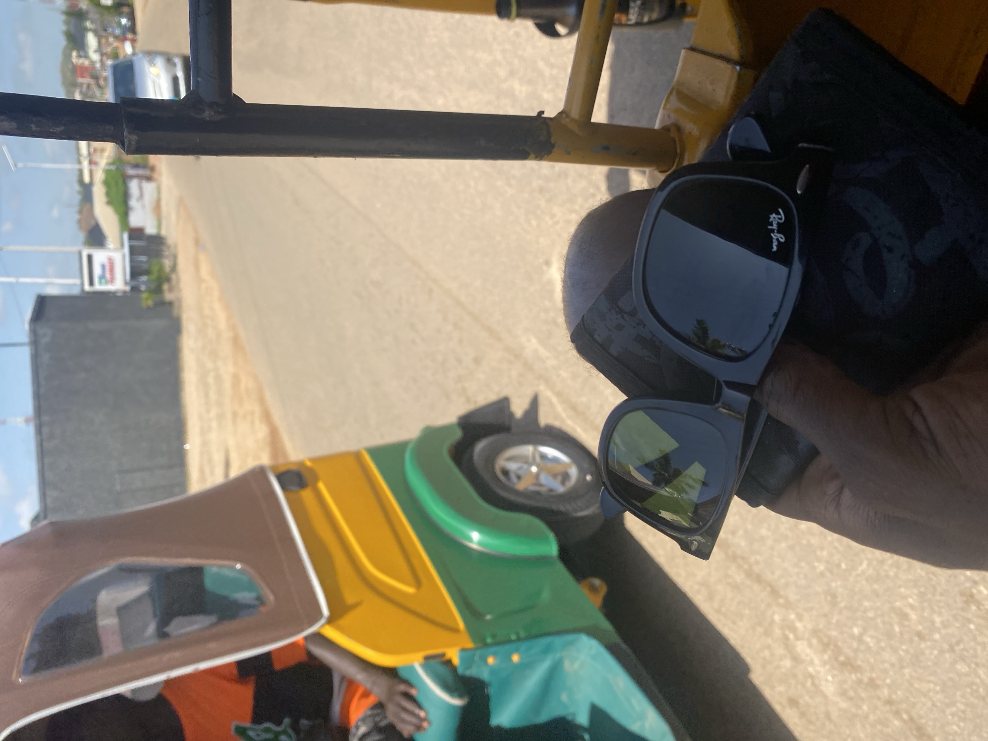 The image captures a first-person perspective from within a motorized rickshaw, commonly known as a "keke napep" in Nigeria. The subject's hand, holding a pair of black Ray-Ban sunglasses, is prominently featured in the foreground. The sunglasses are resting on a dark cloth or bag. Behind the hand, the interior of the rickshaw is visible, including a yellow and green chassis and a portion of a driver's seat occupied by a person wearing an orange shirt with black stripes. The rickshaw is moving on a paved road under a bright, sunny sky with scattered clouds. In the background, a grey building, some smaller structures, a silver car, and distant hills are visible, suggesting an urban or semi-urban environment. This scene is consistent with locations in Gura Top, Nigeria, where keke napeps are a common mode of transportation and the landscape often features such elements.