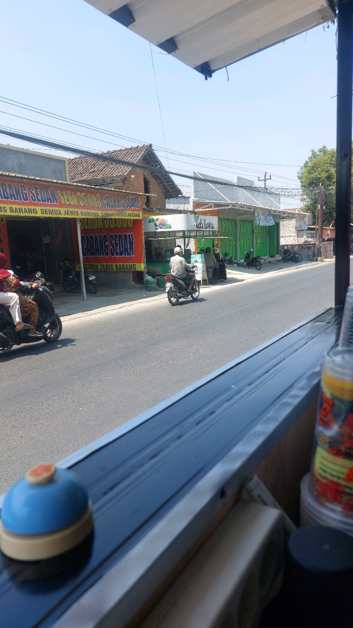 The image captures a street scene in Karas, Indonesia, on a clear, sunny day. The perspective is from a slightly elevated position, looking out from under an awning or overhang.
In the foreground, on a dark blue surface, sits a small, light blue bell with a beige base and an orange button on top. To the right, partially visible, is a clear plastic container filled with red and yellow items, possibly candy or fruit, with a yellow label.
The middle ground features a road with asphalt paving. Several motorcycles are present: one is parked in front of a store, another is being ridden by a person wearing a white shirt and a white cap, moving away from the viewer. Two other motorcycles are parked further down the street. On the left side of the road, two people are riding a scooter, with the passenger wearing a maroon headscarf and patterned clothing.
The background shows a row of shops and buildings. One prominent building has a large red and yellow banner with Indonesian text that appears to advertise a sale. Other shops have green doors and awnings. The sky is clear blue. The overall environment suggests a commercial street in a relatively warm climate.