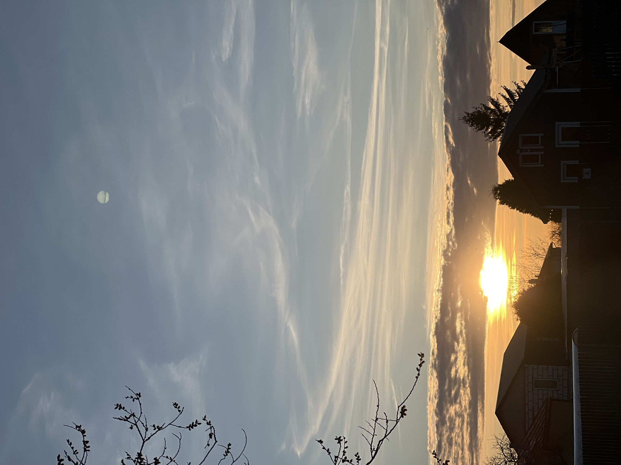 The image depicts a sunset view with a combination of natural and urban elements, likely taken in the city of Riga, Latvia, based on the architectural style of buildings and the visible environment.
**Environment:** The sky dominates the view, featuring a blue gradient that transitions into an orange hue near the horizon where the sun is setting. Wispy clouds are visible across the sky. The image captures the warm and dramatic lighting associated with sunset, casting a golden glow on the clouds and surrounding objects.
**Objects:** Silhouettes of buildings are prominent on the right, partially obscuring the setting sun. These appear to be residential structures with distinct architectural features, including rooflines and window placements. In the foreground, bare branches of a tree or shrub are visible, framing the scene. On the left side of the sky, there is a circular object that could be the moon, or it could be a lens flare or other anomaly.
**Actions/Interactions:** The main action is the sun setting, creating a natural display of light and color. The interplay between light and shadow defines the shapes and details of the buildings and trees.