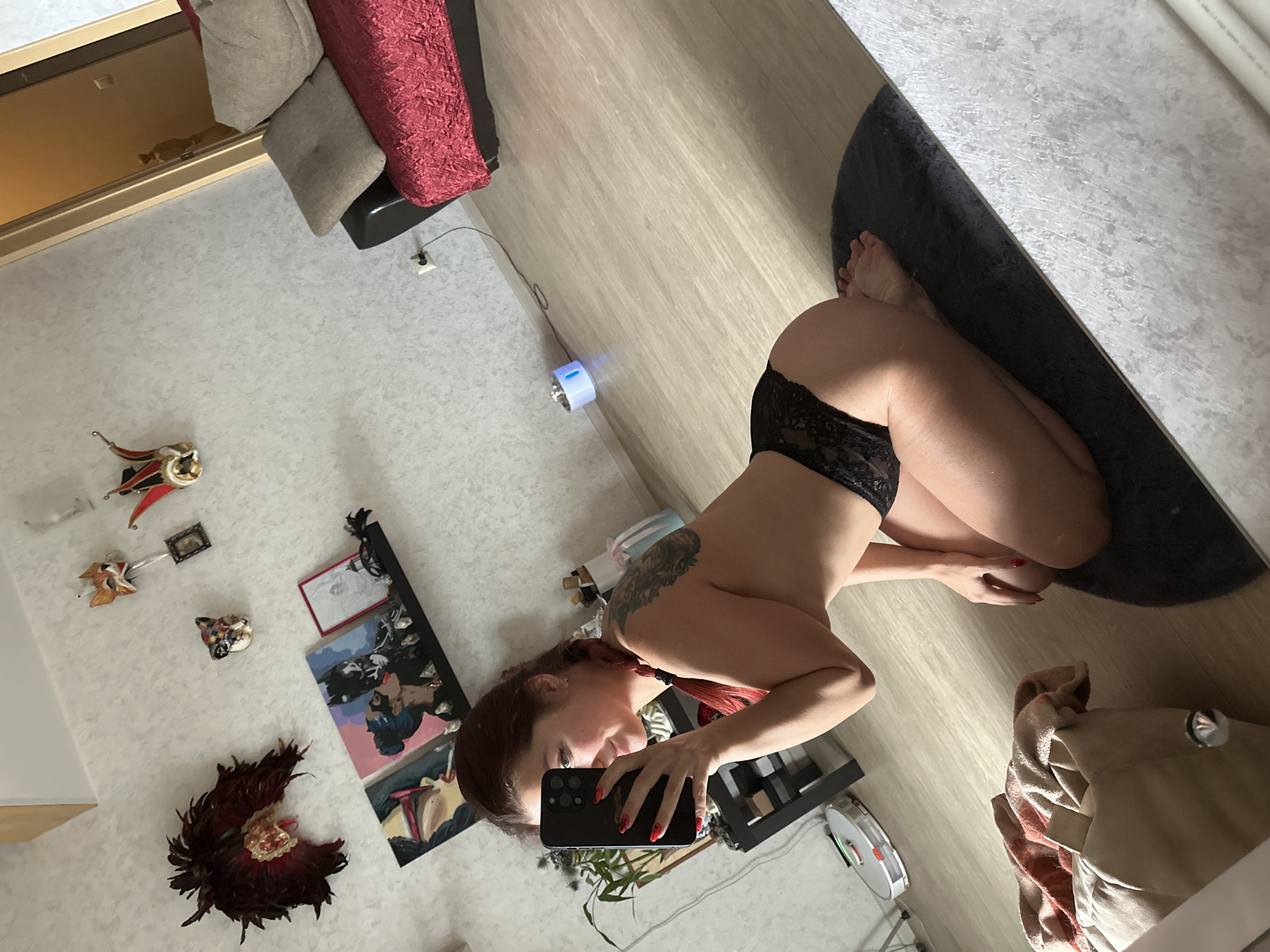 A young woman with red hair in a braid is kneeling on a dark rug in what appears to be a living room. She is holding a black smartphone and taking a mirror selfie, capturing her side profile. She is wearing black lace underwear. The room has light-colored walls with patterned wallpaper. On the wall, there are several decorative masks and three framed paintings, including one depicting Batman and Catwoman. A black shelf is mounted on the wall, holding the paintings and some small decorative items. To the right, a red sofa with two pillows is visible, and beyond that, a doorway leads to another room. In the foreground, a patterned bag or cushion is on the floor. A white robot vacuum cleaner is on the wooden floor near a black side table. A small blue humidifier with a light is also on the floor. The overall impression is of a modern, decorated living space.