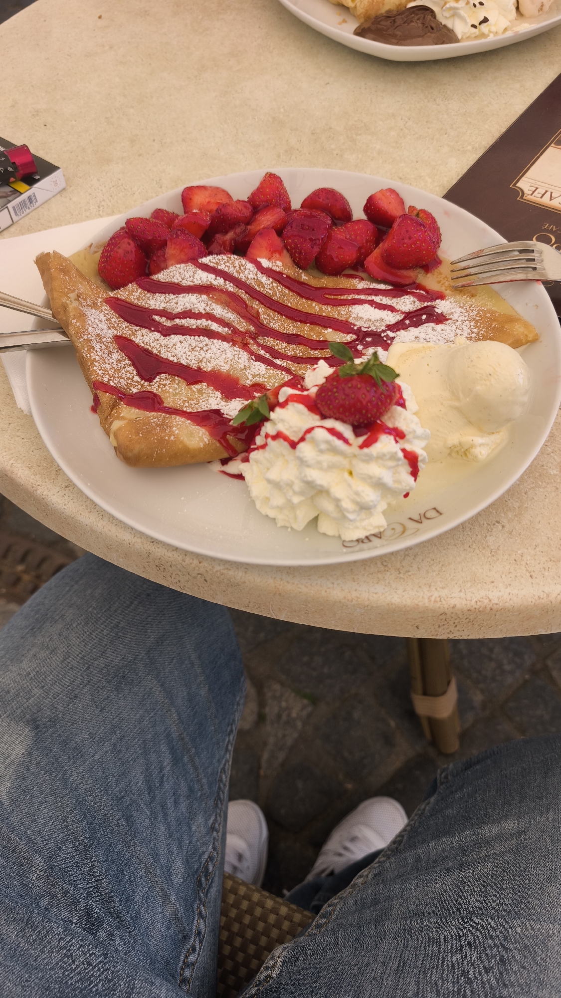 A top-down shot depicts a plate of strawberry crepes topped with whipped cream and a scoop of vanilla ice cream. The crepes are drizzled with a red sauce and dusted with powdered sugar. A fork and knife sit at the edge of the plate. The plate rests on a beige, textured table, with the lower half of a person wearing jeans and white sneakers visible in the foreground. A pack of cigarettes is placed next to the plate. The scene suggests an outdoor cafe setting.