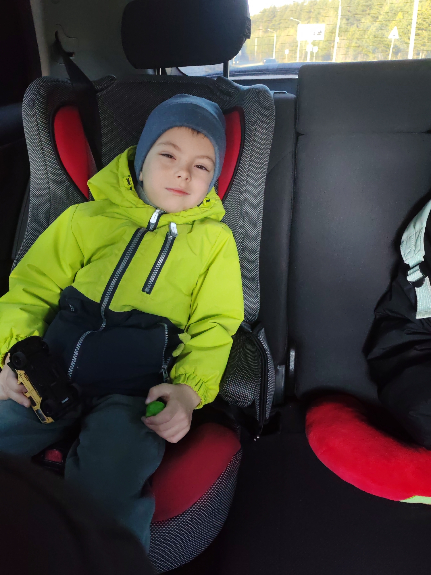A young boy, wearing a blue hat and a bright yellow jacket with a dark blue bottom half, sits in a car seat in the backseat of a car. He is holding a toy car in his left hand and a small green ball in his right. The car is moving, as suggested by the blurred background visible through the rear window which includes trees and road infrastructure. The backdrop indicates the location is Chaikovsky, Russia. A red cushion or pad is partially visible next to the boy.