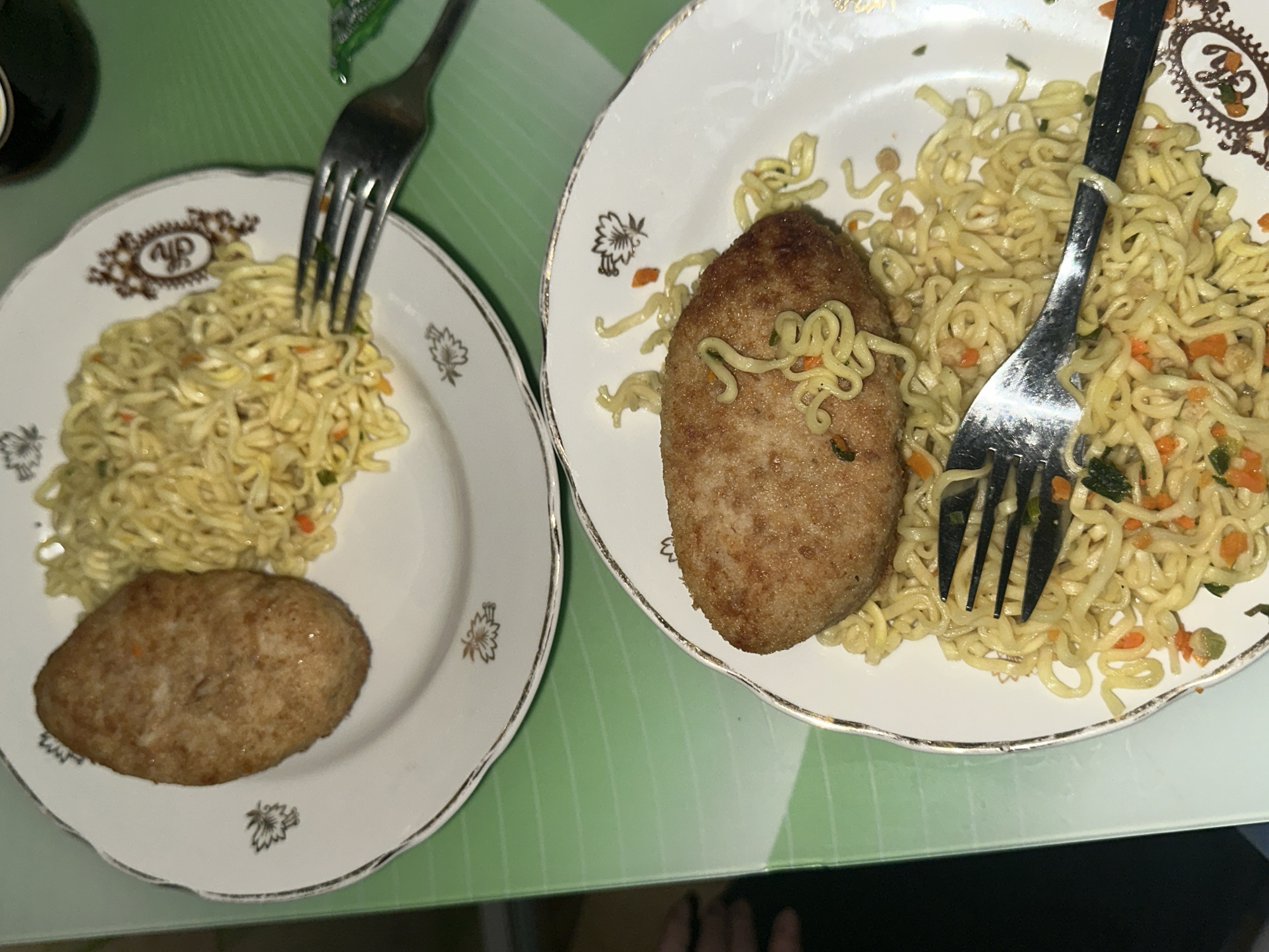 Two plates of food are positioned on a light green surface. Each plate contains a fried cutlet and a portion of instant noodles, with forks placed on top of the food. The plates feature a white background with ornate gold-colored designs around the edges. The location and city are not discernable from the image.