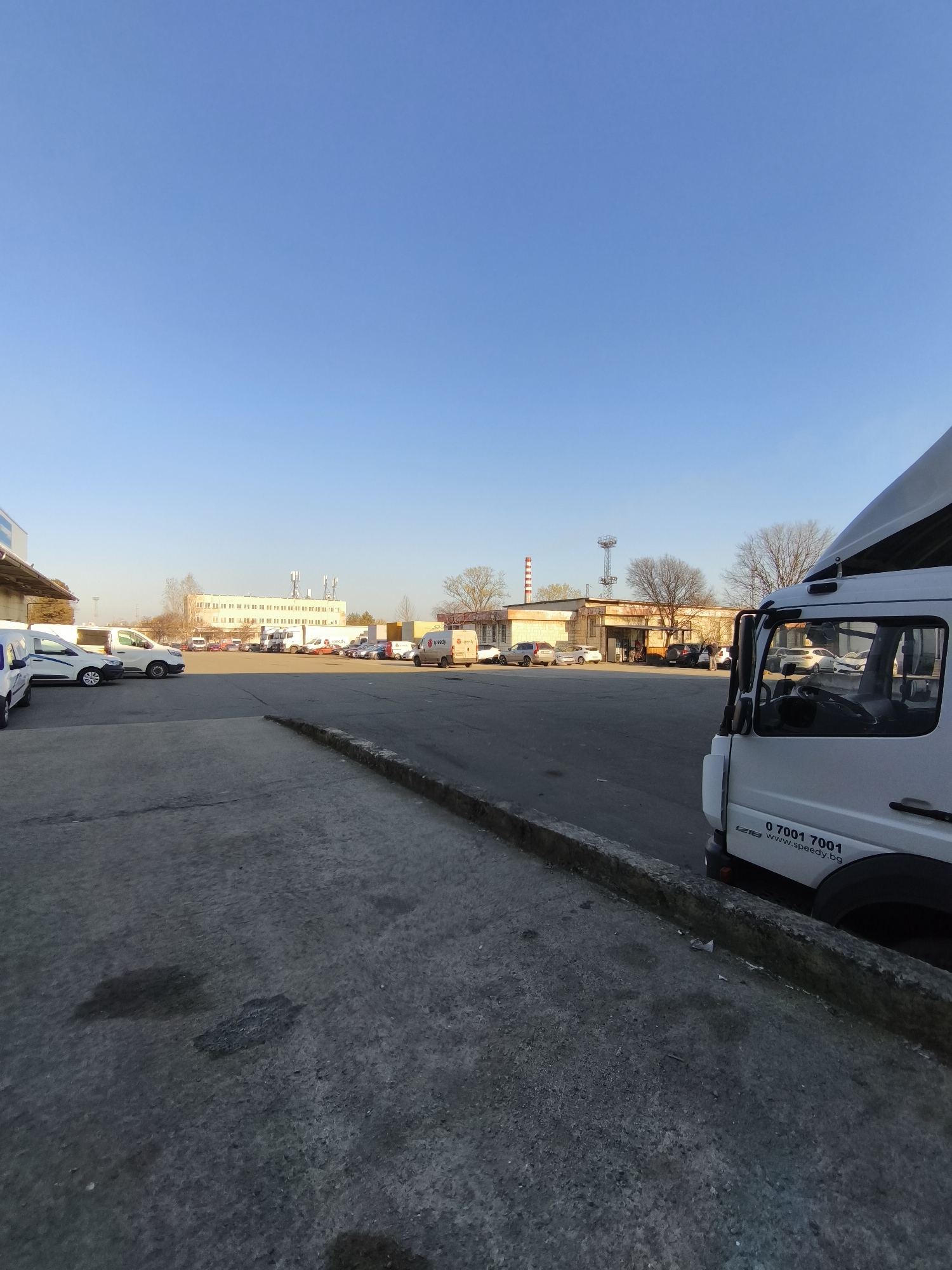 The image depicts an outdoor parking lot scene in what appears to be an industrial or commercial area. The sky is clear and blue. The foreground shows a concrete surface leading to a curb. A white van or truck is partially visible on the right side of the image, bearing the number "0 7001 7001" and the website "www.speedy.bg". In the background, several cars and vans are parked in a lot, and various buildings are visible. The presence of Speedy-branded vehicles indicates that this is likely a Speedy logistics center or depot. The location is Burgas, Bulgaria. There are no people visible in the image.
