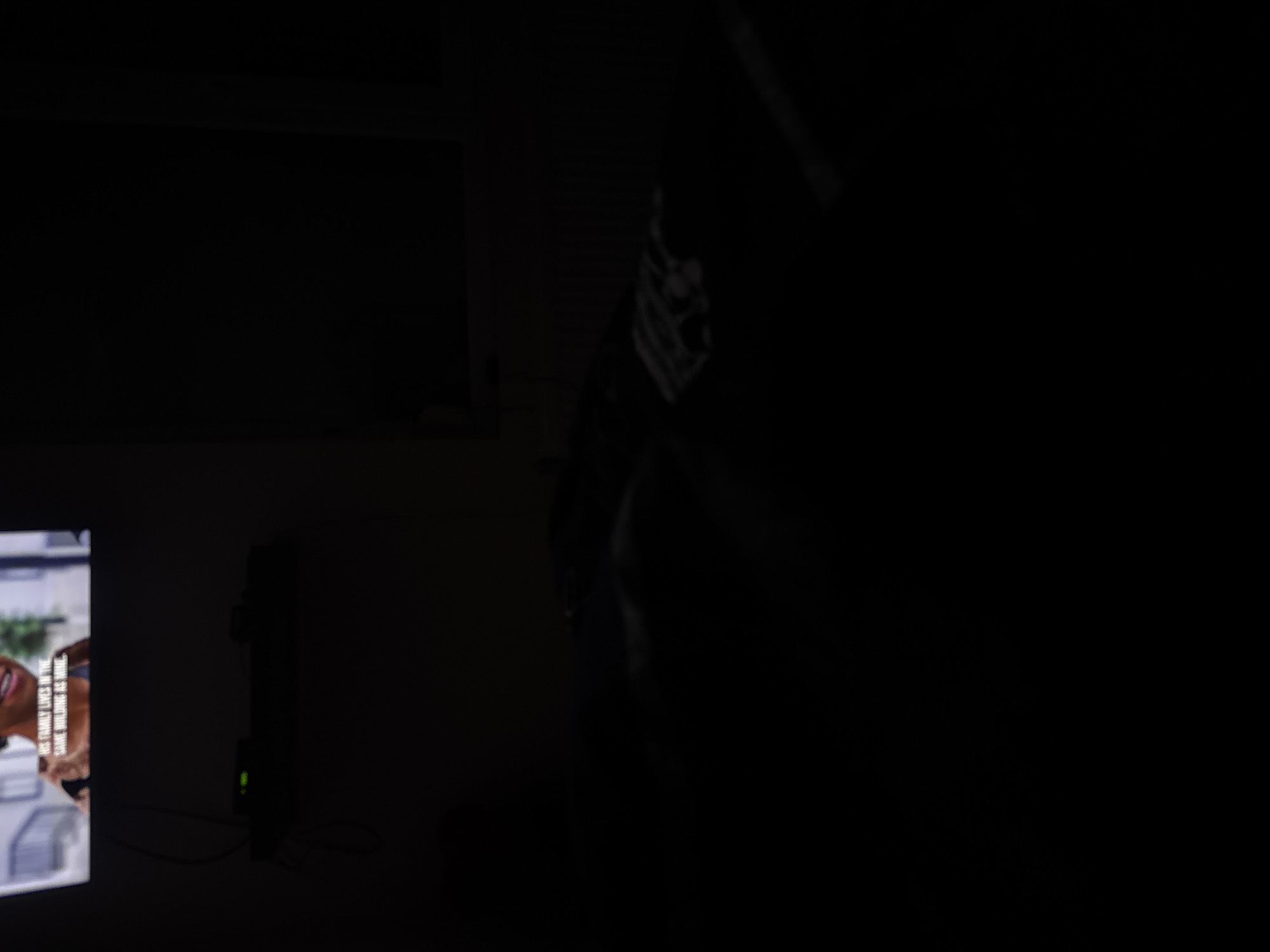 The image shows a dark room with a television in the lower left corner displaying a scene with a woman and text. The text reads "HIS FAMILY LIVES IN THE SAME BUILDING AS MINE". In the center of the image, there is a tall, blurred figure that appears to be a person holding a long object. The environment is dark, making it difficult to discern details. The location is not discernible from the image.