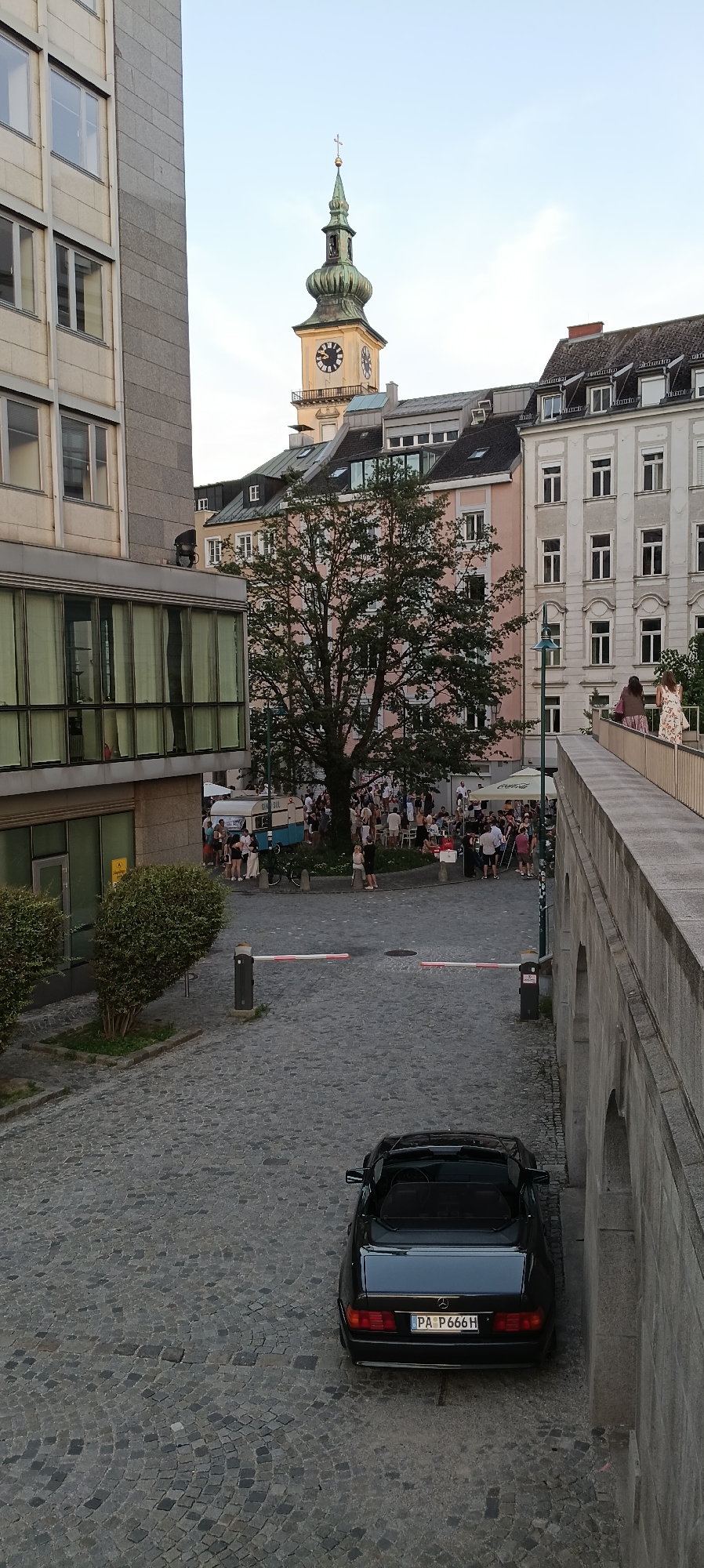 The image depicts a scene in Linz, Austria, likely near the Hauptplatz (Main Square), given the prominent church tower with a clock. In the foreground, a black convertible car with the license plate "PA P666H" is parked on a cobblestone street. The street is partially blocked by a white and red barrier. To the left, a modern building with large windows and green curtains is visible, with manicured bushes in front of it.
In the middle ground, a large tree with lush green foliage stands in the center of a gathering of people. The crowd appears to be enjoying an outdoor event or market, with some seated at tables under umbrellas. Several buildings with traditional European architecture, featuring pitched roofs and multiple windows, surround the gathering. One building is painted a pale pink, another light gray, and a third has a dark gray roof.
In the background, the distinct steeple of a church tower, featuring a green onion dome and a cross, rises above the buildings. A clock face is clearly visible on the tower. To the right of the image, a stone walkway with arched structures runs along the side, suggesting it's an elevated pathway or bridge. Two women are walking on this pathway, one wearing a floral dress and the other a brown top and patterned skirt. The sky is a clear, pale blue, indicating daytime.