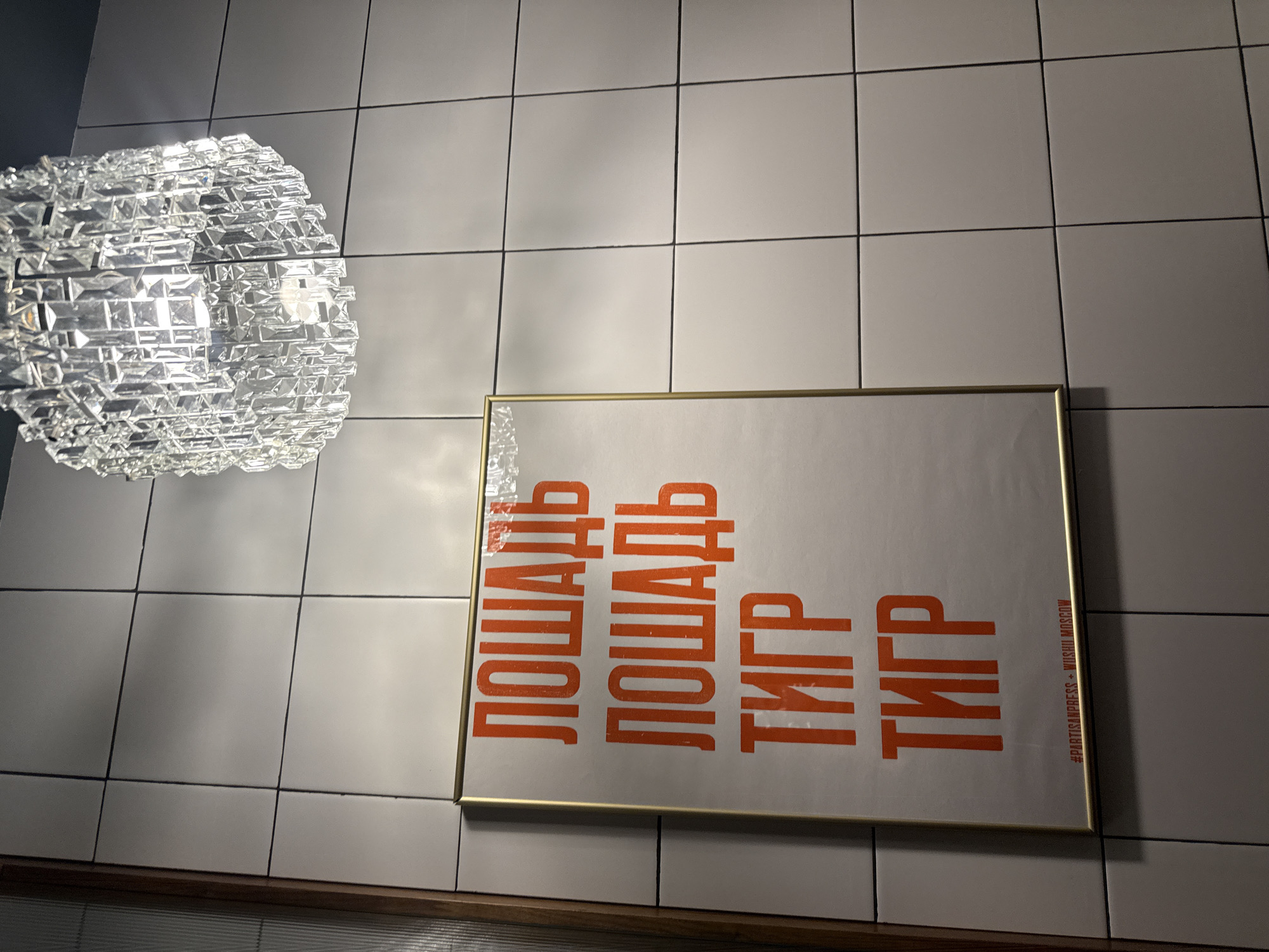 The image displays an interior scene focusing on a textured wall and a decorative light fixture. The wall is covered in light-colored, possibly white or light gray, square tiles with dark grout lines, suggesting a modern or minimalist aesthetic.
In the upper left quadrant, a large, spherical chandelier hangs. It is intricately designed with numerous clear, faceted glass or crystal pieces, which reflect and refract the internal light, creating a sparkling effect. The light source within the fixture is visible through the crystalline structure.
On the tiled wall, slightly to the right and lower than the chandelier, hangs a rectangular poster in a thin, golden frame. The poster features bold, orange Cyrillic text. The words "ЛОШАДЬ" (Loshad', meaning "Horse") are printed twice, one above the other, followed by "ТИГР" (Tigr, meaning "Tiger") also printed twice. Along the right edge of the poster, smaller, vertically oriented text reads "#PARTISANPRESS + WIJSHU MOSCOW," providing a location cue for Moscow, Russia.
Below the tiled wall and poster, a dark wooden ledge or shelf is visible, running horizontally across the bottom of the frame. The lighting in the scene appears to be artificial and bright, highlighting the details of the chandelier and the poster. The image captures a static view of interior decor, likely in a commercial or public space.