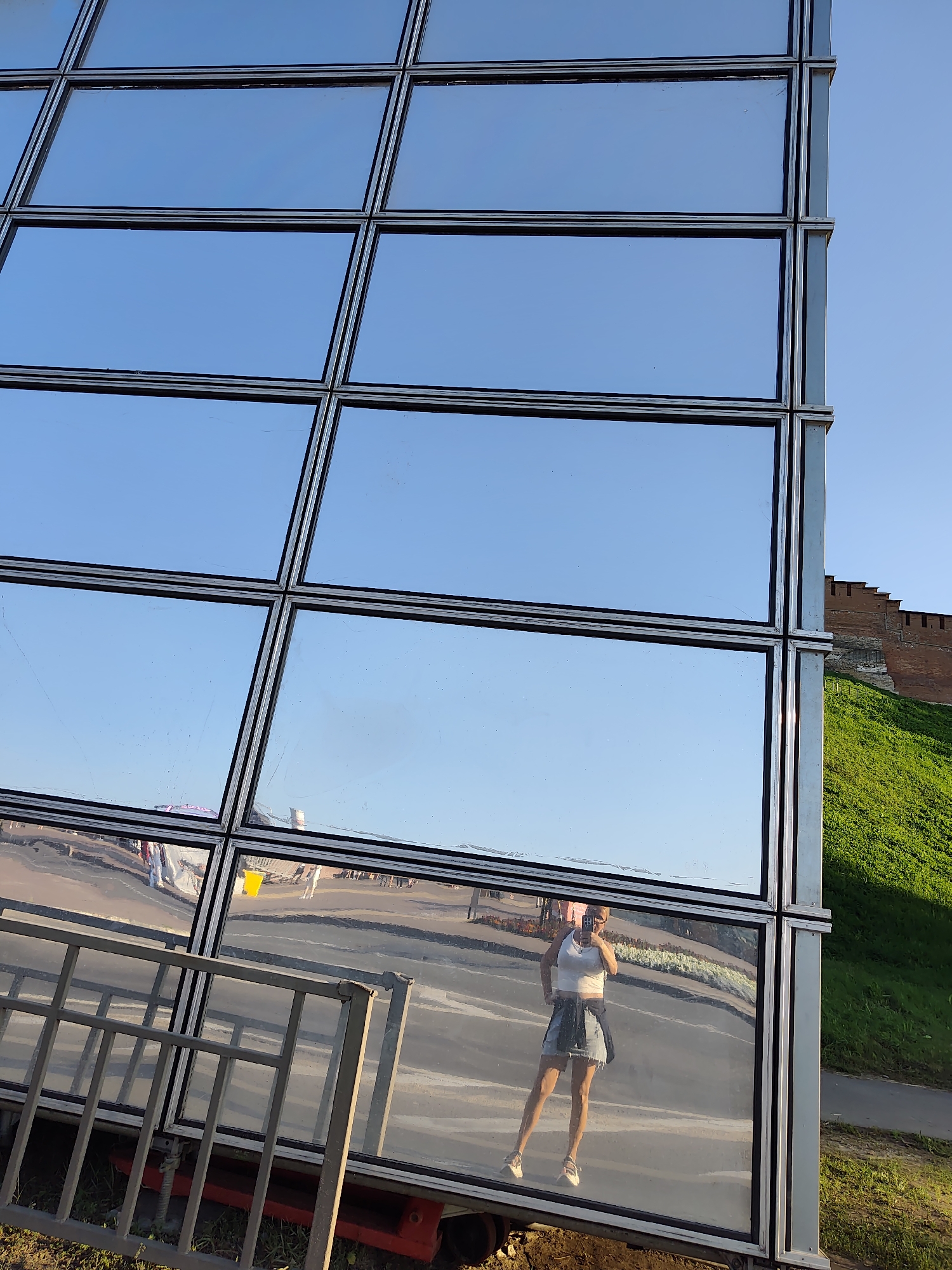 The image captures a reflection in a modern glass building, showcasing a woman taking a selfie. She is wearing a white tank top, a denim skirt, and sneakers, with a jacket tied around her waist. Her reflection is visible in the lower half of the building's facade, which is composed of numerous rectangular glass panes. The building is situated outdoors, with a clear blue sky above. To the right of the building, a lush green hill slopes upwards, topped by an ancient red brick fortress wall, characteristic of the Nizhny Novgorod Kremlin. The reflection also shows a paved area with some parked vehicles and people walking in the background, suggesting an urban setting. The foreground includes a metal fence and some ground cover.