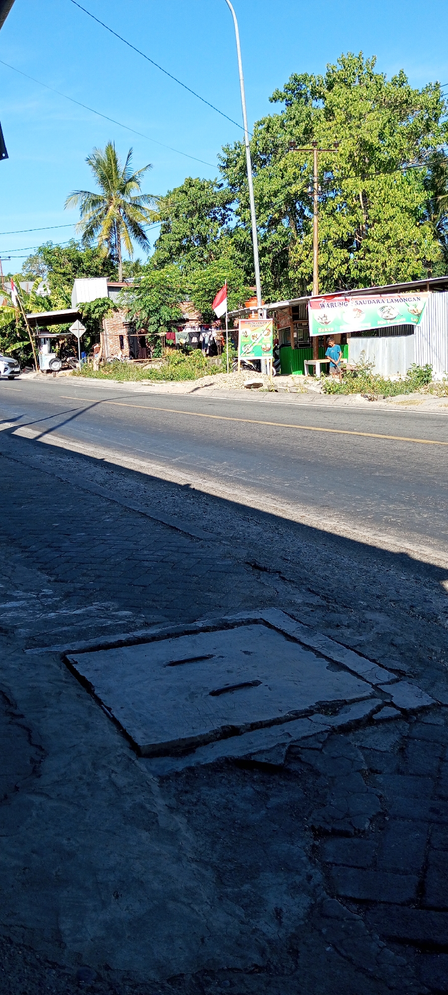 The image depicts a street scene in Pakkanna, Indonesia, on a bright, sunny day with a clear blue sky. The foreground shows a concrete storm drain cover on a cracked, textured pavement. A paved road with a white line marking runs through the middle ground, casting a shadow from an unseen object. On the right side of the road, there is a roadside establishment with a corrugated metal roof and a green awning. A sign above the awning reads "WARUNG SAUDARA LAMONGAN" with pictures of food items. A young person wearing a blue t-shirt and dark pants is standing in front of the establishment, holding a broom. To the left of the establishment, a red and white Indonesian flag is visible. Further to the left, there is another small building with laundry hanging out to dry. A white car is parked on the side of the road near this building. A tall palm tree and lush green foliage are visible behind the buildings. A lamppost stands tall on the right side of the image, extending into the sky. Electrical wires crisscross the sky above the scene.
