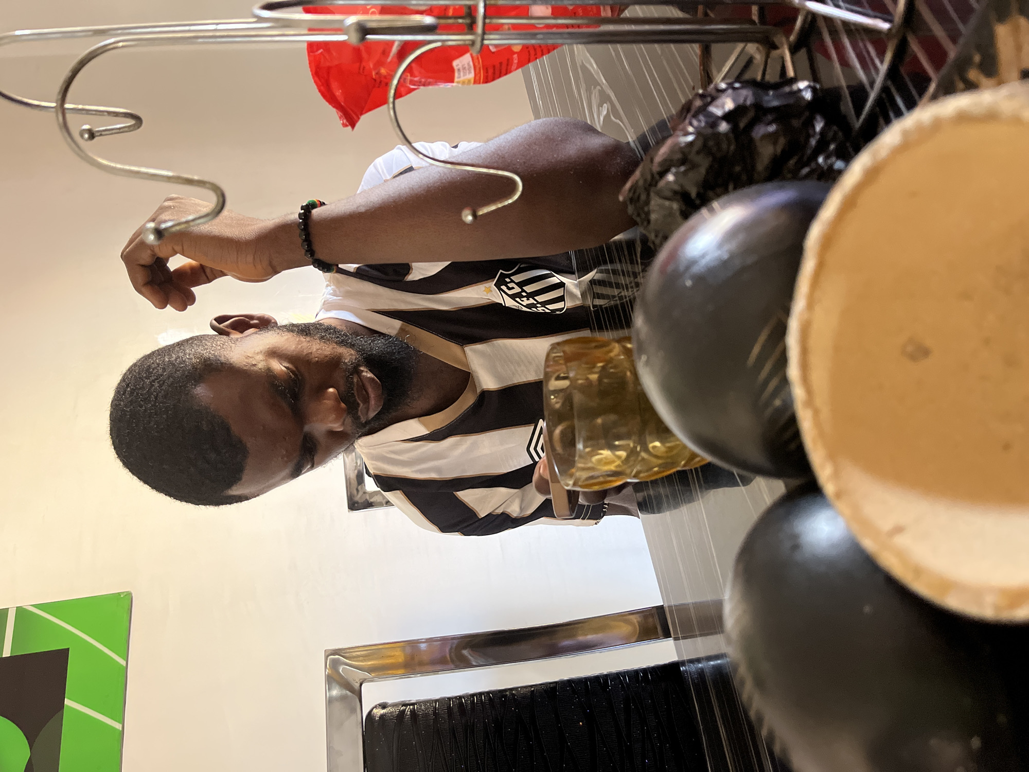 A man with dark hair, a trimmed beard, and a black and white striped shirt is seen holding a glass of amber-colored liquid at a location in Jos, Nigeria. He is wearing a beaded bracelet. The man is sitting at a reflective black surface. In the foreground, partially obscuring the scene, are several round, black objects and a hollow, light-colored container. A metal rack hangs above the man's head with a red package visible. The background consists of a white wall with a green decorative element and a chrome-plated frame. The scene is indoors, possibly a kitchen or dining area.