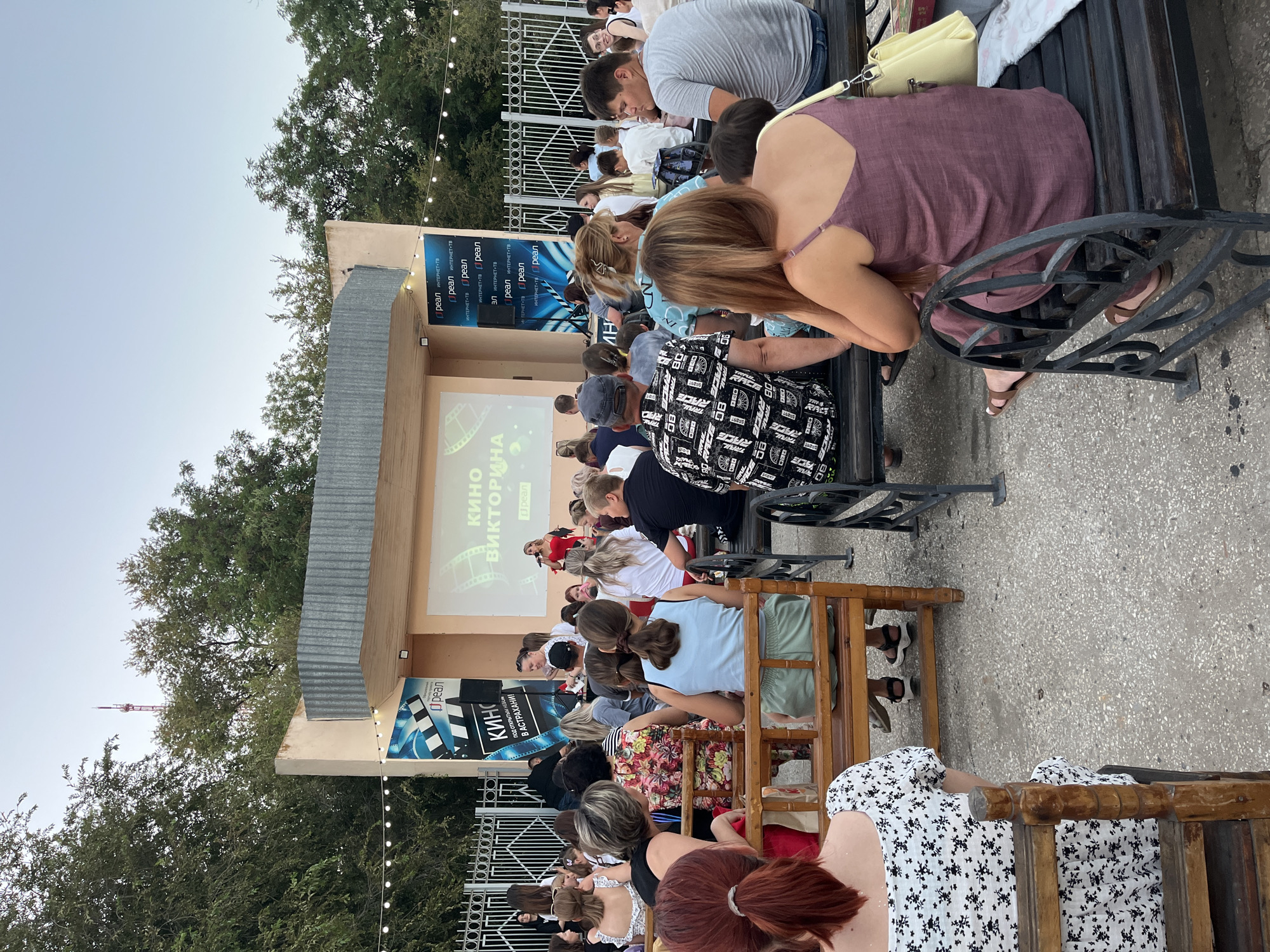 The image depicts an outdoor movie screening at the Astrakhan State Theatre of Opera and Ballet, located in Astrakhan, Russia. A diverse crowd of approximately fifty people is seated on benches, facing a large screen. The screen displays text in Russian, "КИНО ВИКТОРИНА," which translates to "Movie Quiz." A woman in a red dress stands near the screen, holding a microphone, suggesting she is hosting or presenting. The setting includes trees in the background and decorative string lights overhead, indicating an evening event. The atmosphere appears relaxed and engaged, with attendees of various ages enjoying the open-air cinema experience.