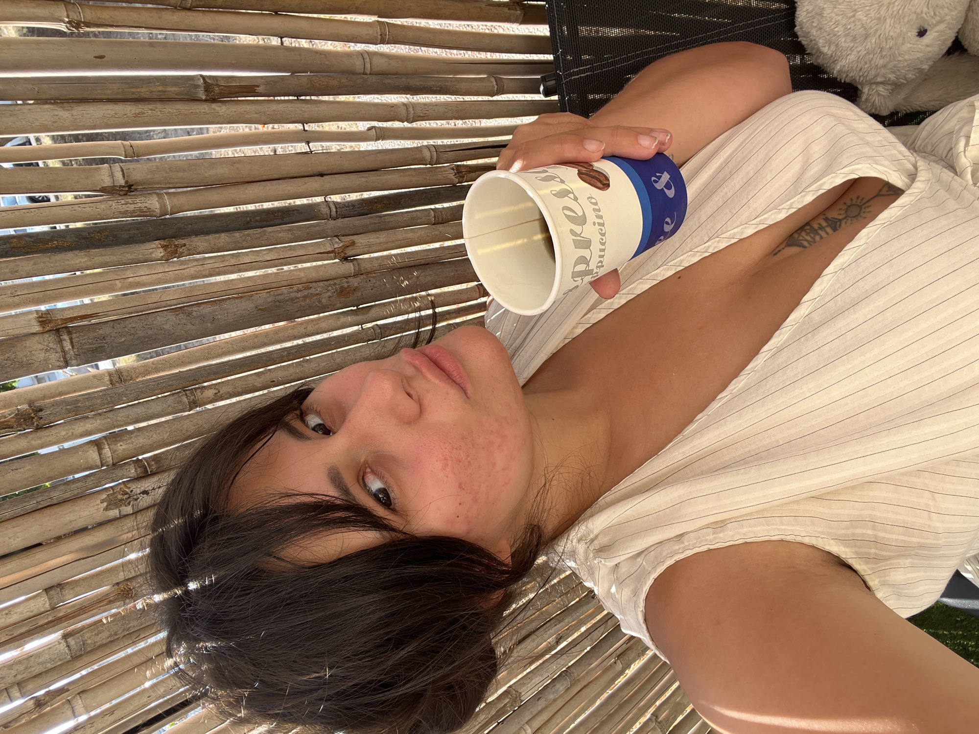 A young woman with dark hair pulled back and bangs is sitting outdoors, against a backdrop of vertical bamboo stalks. She is holding a white paper coffee cup with blue and brown text that reads "Espresso Cappuccino" in her right hand. She is wearing a light-colored, sleeveless, pinstriped top with a deep V-neckline, revealing a tattoo on her chest. Her left arm is visible, and she has some light body hair under her arm. The lighting suggests it is daytime, with sunlight casting shadows. The environment appears to be a casual outdoor setting, potentially a cafe or patio. No specific landmarks in Kiryat Yam, Israel, are identifiable in the image.