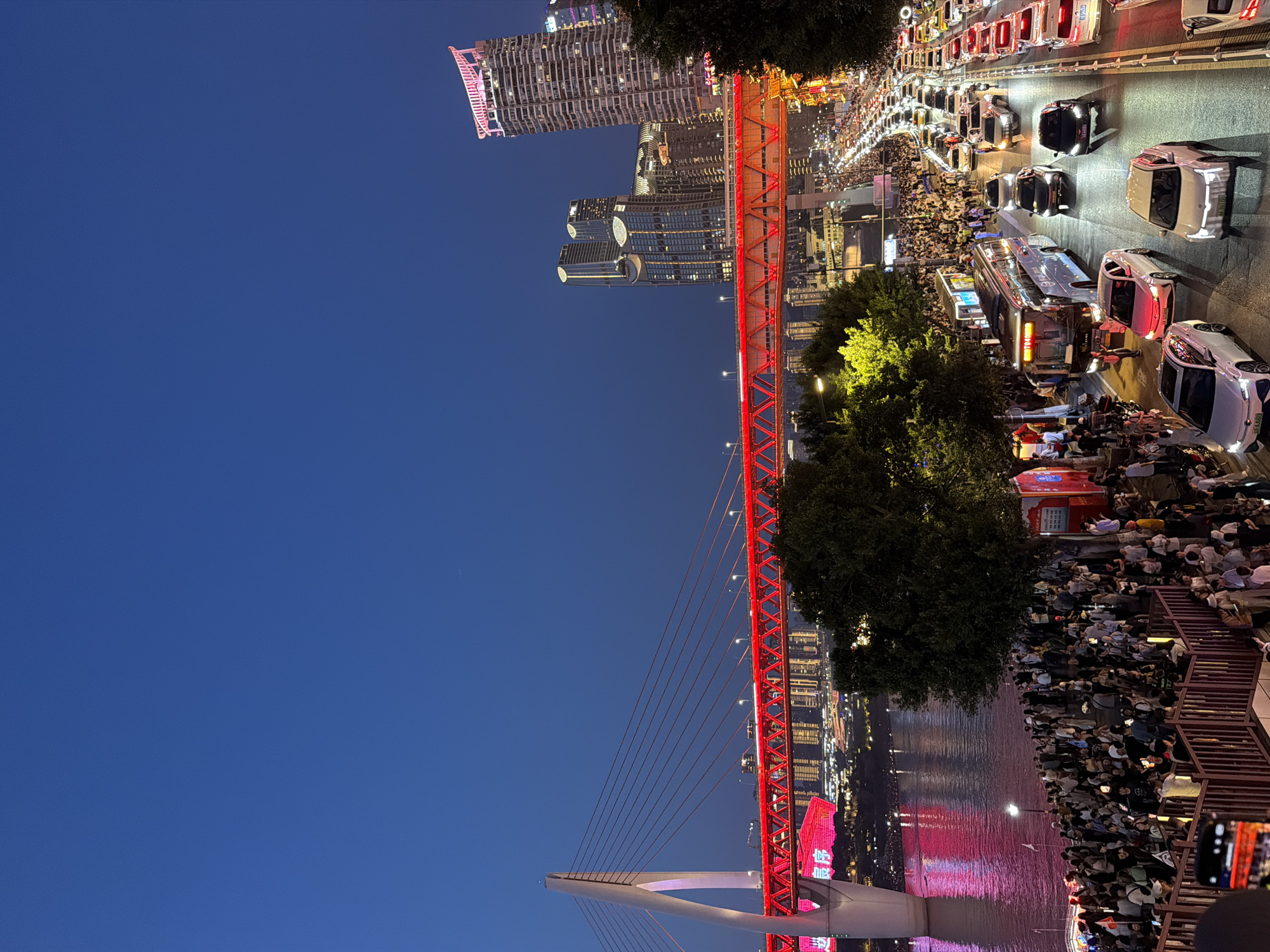 The image depicts an evening scene at the Yuzhong District in China, likely at dusk or early night. A large, illuminated red suspension bridge spans across a body of water, reflecting the vibrant lights. On the riverbank and elevated walkways, a significant crowd of people is gathered, observing the illuminated cityscape and bridge. The background features a cluster of modern skyscrapers, also lit up, contributing to the urban ambiance. In the foreground, a busy road is filled with a steady stream of vehicles, including cars and a bus, with their headlights and taillights creating streaks of light. Trees with illuminated leaves and the glow of streetlights dot the scene, adding to the lively atmosphere. The sky is a deep, dark blue, indicative of the late hour.