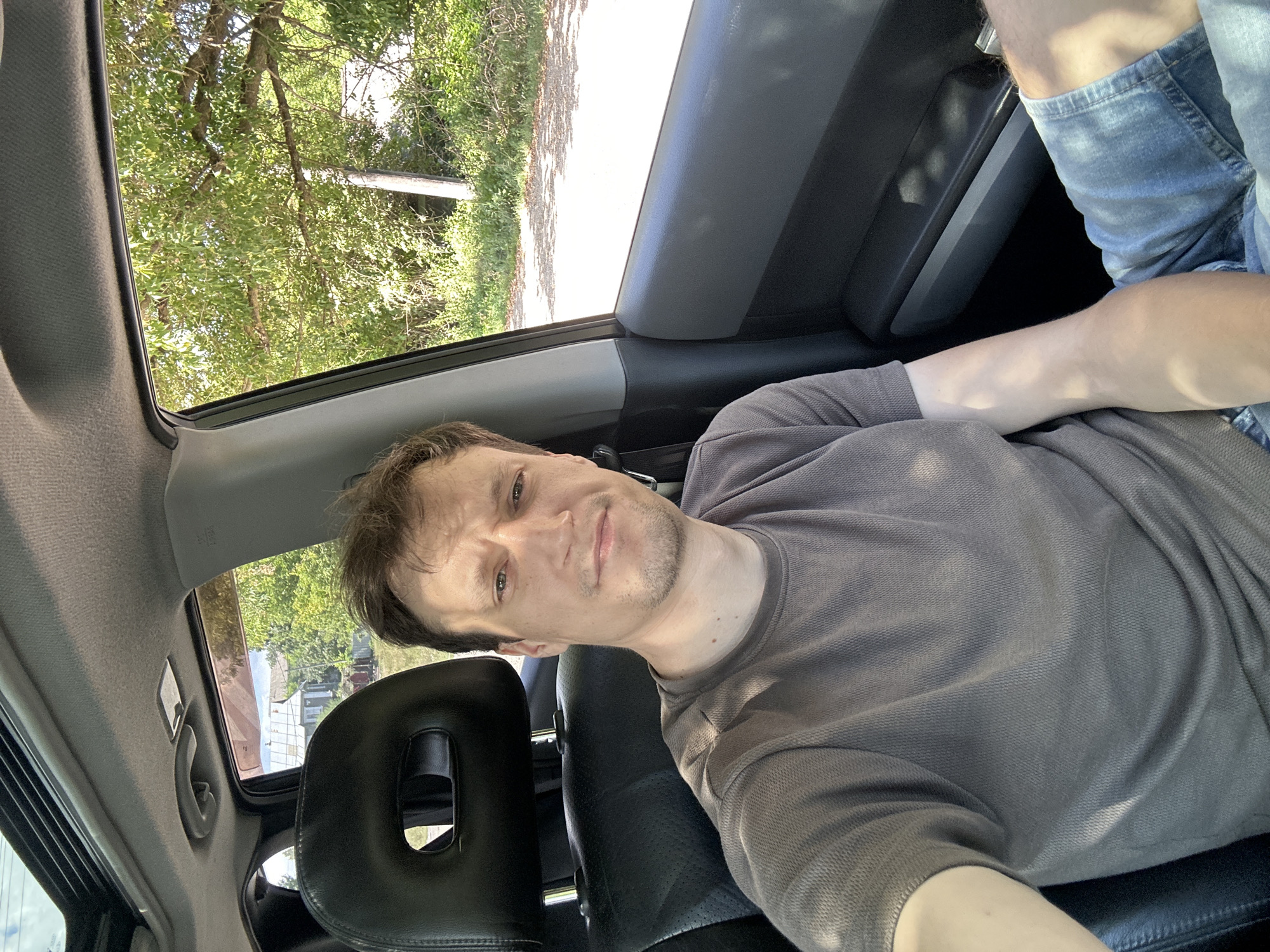 A fair-skinned male with brown hair and stubble is sitting in the driver's seat of a car, taking a selfie. He is wearing a short-sleeved, gray, textured t-shirt and denim shorts. The car has a black leather headrest with an oval cutout. Outside the car window, lush green trees and a building are visible under a bright, sunny sky. The overall setting appears to be a rural or suburban area, potentially in or around Makiv, Ukraine, given the presence of trees, a simple building, and a paved road.
