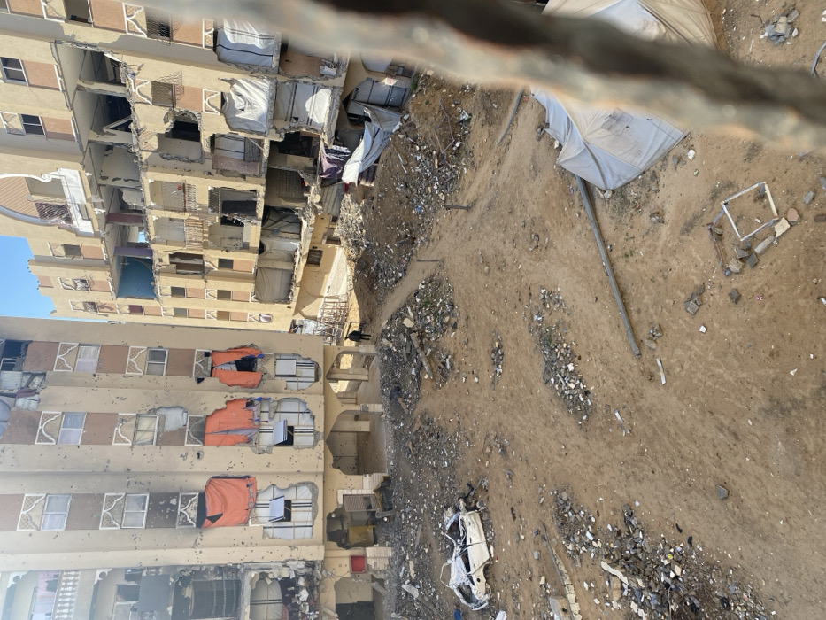 The image depicts a heavily damaged urban area in Khan Yunis, Palestinian Territory, viewed from an elevated perspective. Two multi-story buildings dominate the background, both showing extensive structural compromise. The building on the left, a light tan color with decorative window frames, features numerous circular impact marks across its facade and multiple blown-out windows, some covered with torn orange tarps. The lower levels exhibit open archways and visible rubble. The building on the right, also light tan, is more severely damaged, with large sections of walls missing, exposing interior rooms, utility lines, and collapsed balconies. White tarps cover several openings on this structure.
The foreground and mid-ground consist of an unpaved, sandy-brown ground covered with significant amounts of rubble, debris, and scattered fragments of concrete, metal, and plastic. Mounds of dirt and debris are prominent, with narrow dirt paths winding through them. On the left side of the foreground, the heavily damaged, white chassis of a vehicle, missing its roof and most body panels, rests on the ground amidst debris. In the lower right, a collapsed light-colored tent or tarp structure and a segment of a fallen pole or pipe are visible.
One individual, dressed in dark clothing, is visible walking on a dirt path in the mid-ground, near the base of the left building. The sky above is clear and light blue. A blurry, dark, vertical metal object partially obstructs the view on the far right of the foreground, suggesting the camera's position behind a railing or similar structure.