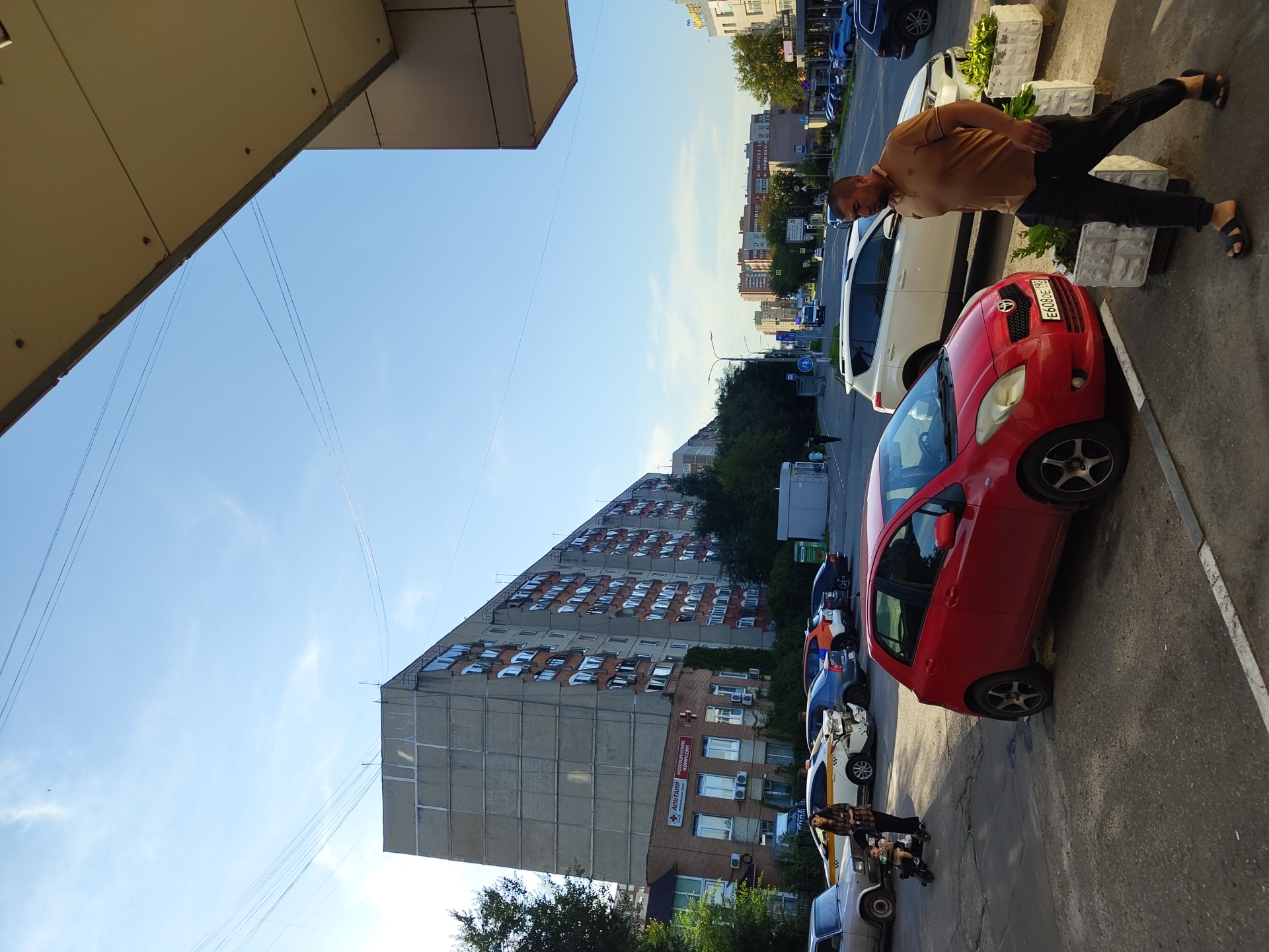 In Zhukovsky, Russia, on a clear day with a blue sky and scattered clouds, a street scene unfolds. A large, grey, multi-story apartment building dominates the background, its facade showing rows of windows and balconies. In front of it, a smaller brick building houses a medical clinic, identified by signage.
The foreground features a paved street and sidewalk area. Several vehicles are parked along the street. A red hatchback is prominently positioned in the lower right quadrant of the image. To its left, a white SUV is parked, and further back, a yellow taxi with visible damage to its rear bumper is also parked. A woman is seen with a child in a stroller near the taxi.
A man in a brown polo shirt and black pants is walking in the foreground, heading towards the right side of the frame. He is wearing dark sandals. The environment includes streetlights and some greenery. The overall impression is that of a typical urban residential and commercial area in the afternoon.