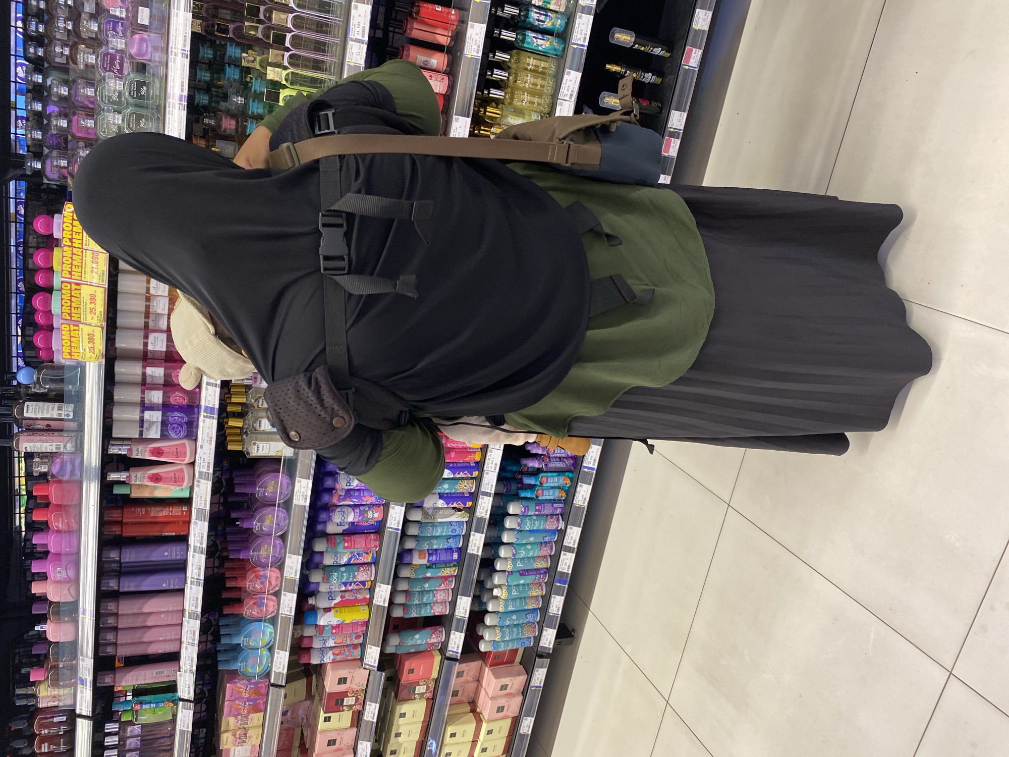 A person wearing a black hijab and a green top is standing in a supermarket aisle, facing away from the camera. They are carrying a baby in a black baby carrier on their back. The shelves behind them are stocked with various perfumes and personal care products, displayed in pink, purple, and clear bottles. The floor is tiled with light-colored squares. The lighting appears to be artificial, typical of an indoor retail environment. The scene is likely a commercial setting where personal care products are sold, such as a pharmacy or department store within Banda Aceh, Indonesia.
