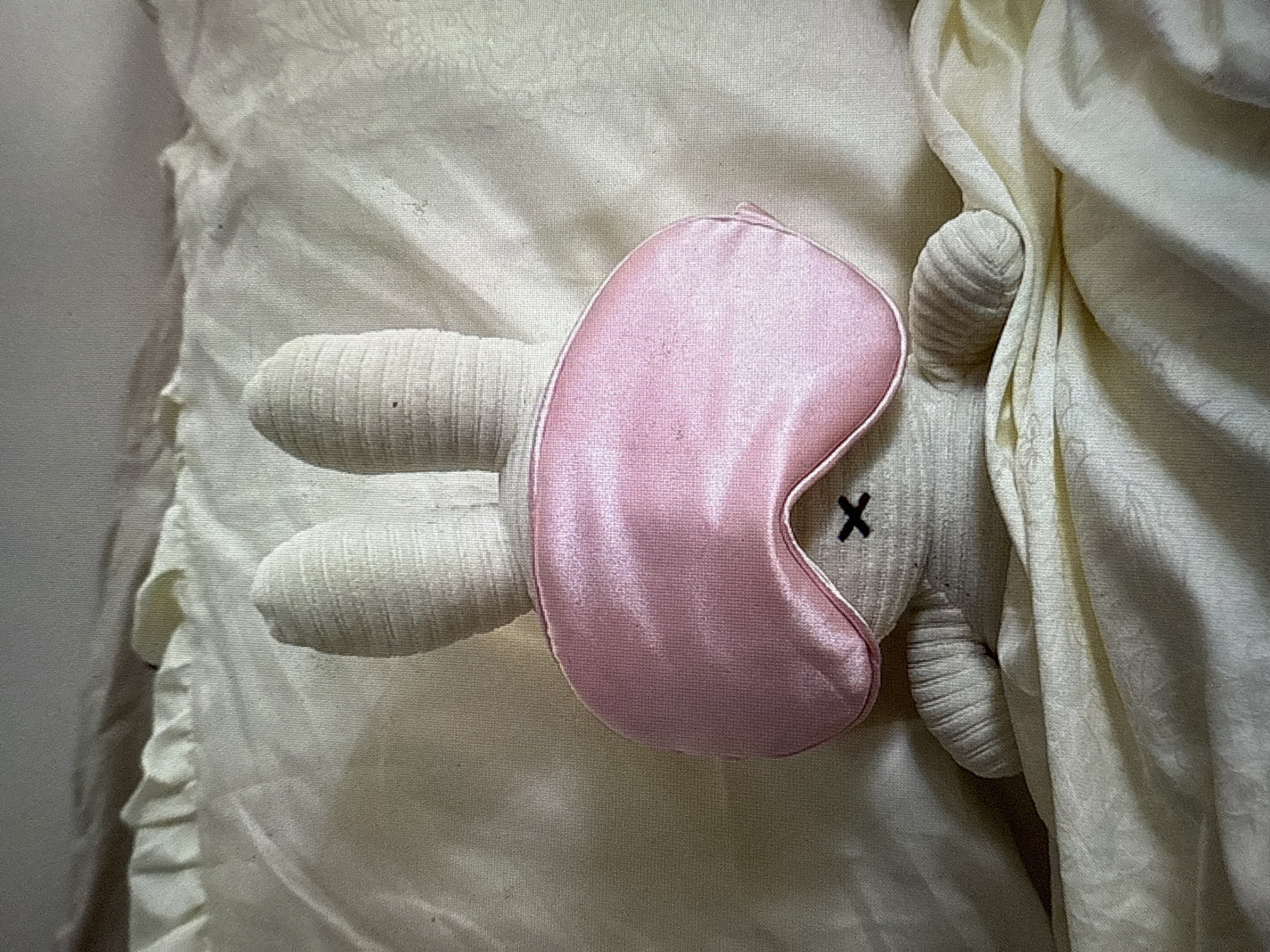 A close-up shot depicts a stuffed rabbit toy with a pink sleep mask covering its eyes, lying on a bed. The rabbit is made of a cream-colored, ribbed fabric, and its eyes are marked with black "X" stitches. The bed has cream-colored sheets with subtle patterns. The lighting appears soft and diffused. The location is unspecified.