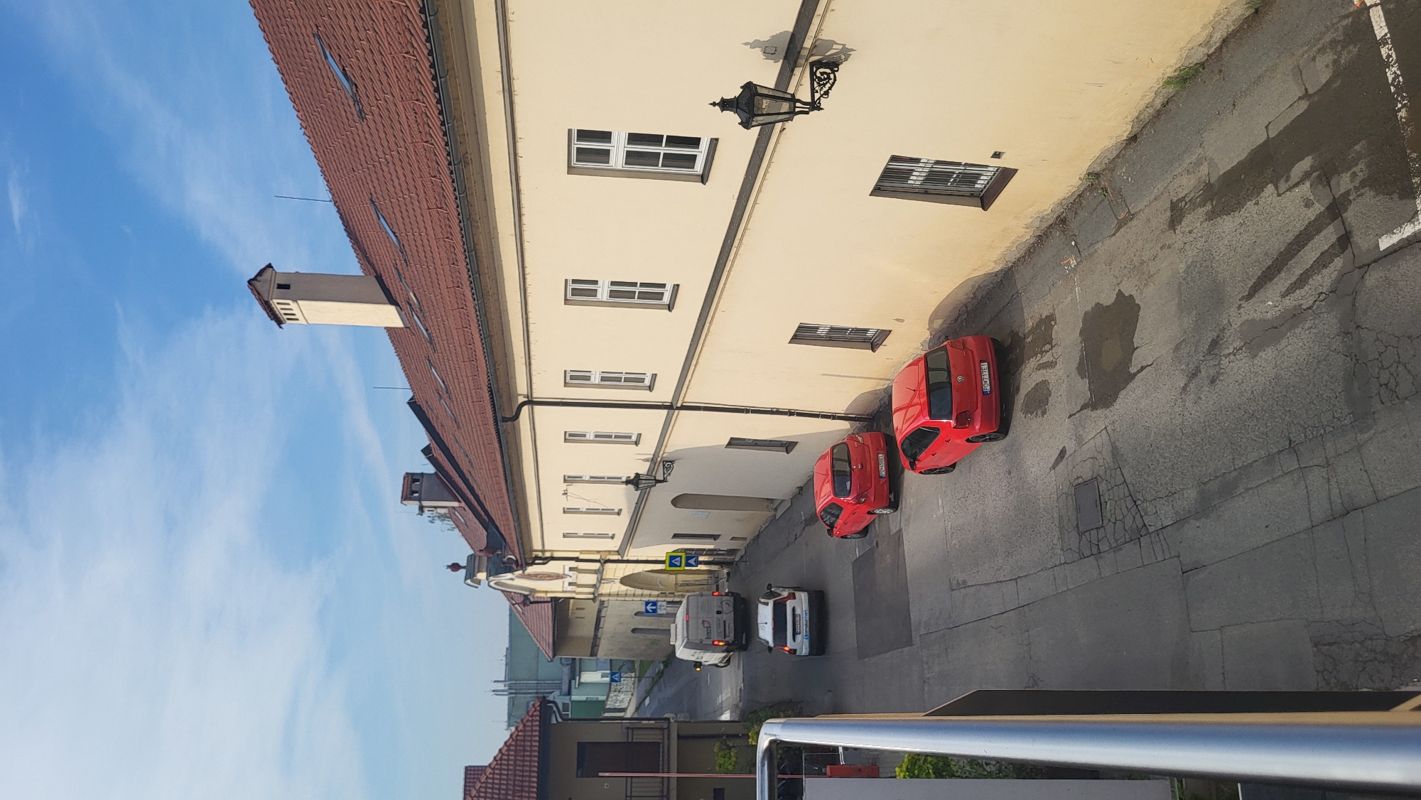 The image shows a street scene in Prešov, Slovakia, under a clear blue sky with scattered clouds. The street is lined with buildings, including a prominent, long, cream-colored building with a red tile roof and multiple windows. The building has a historical architectural style, indicated by its facade and a large archway visible in the distance.
Several vehicles are present on the street. In the foreground, a red car is parked, followed by another red car also parked on the side of the road. Further up the street, a white SUV is parked, and behind it, a white van is driving. The road surface is asphalt with visible cracks and some damp patches, suggesting recent rain.
Architectural elements include chimneys on the roof of the cream-colored building, and decorative lanterns mounted on the exterior walls. The archway in the distance appears to be an entrance to a complex or historical site, with blue and yellow directional signs visible. The scene depicts a quiet street in a city, possibly in a historical district, during daylight hours. There are no people visible in the image.