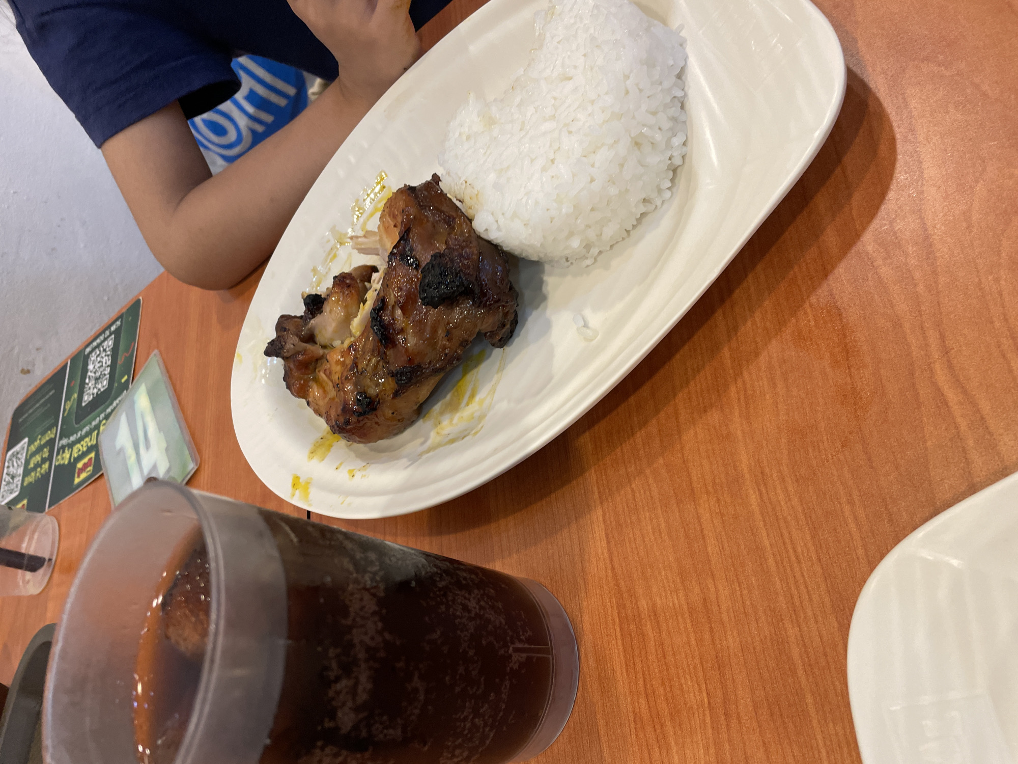 An overhead, close-up shot captures a meal served on an orange-brown wooden table inside a casual dining restaurant. The primary subject is a white rectangular plate holding a portion of grilled chicken, characterized by its charred skin and a glossy, golden-brown glaze, accompanied by a generous mound of white, steamed rice. Yellowish drippings are visible on the plate beneath the chicken. To the left, a clear plastic cup filled with a dark, effervescent carbonated drink is present. In the background, a person's forearm and upper arm, clad in a dark blue t-shirt, rest on the table. Behind the person, a blue and white logo or branding, resembling that of "Mang Inasal," is partially visible on the wall or a display. Other table items include a green placemat or promotional sheet featuring QR codes and text, and a small green-framed sign displaying the number "14". The scene depicts a typical meal experience at a Mang Inasal restaurant in Davao City, Philippines.
