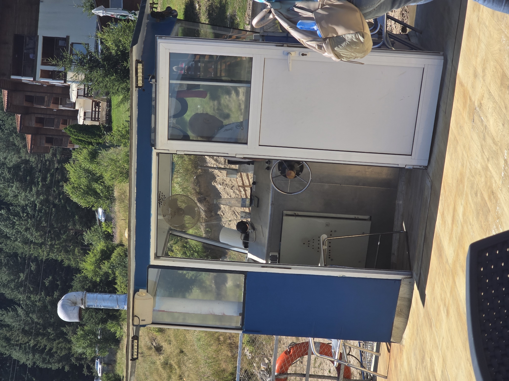 The image depicts a close-up view of the control cabin of a ferry or similar vessel docked on the shore of Sarnitsa, Bulgaria. The cabin is constructed of blue and white panels and glass. Inside, a person, seen from the side, is seated at a control panel, with a steering wheel visible in front of them. A fan is mounted on the wall behind the controls. Outside the cabin, on the deck of the vessel, a life preserver is visible, along with chairs. In the background, the lush green trees of a forest rise up a hillside, and buildings with wooden facades, characteristic of Sarnitsa, are nestled amongst the trees. The scene appears to be set on a sunny day.