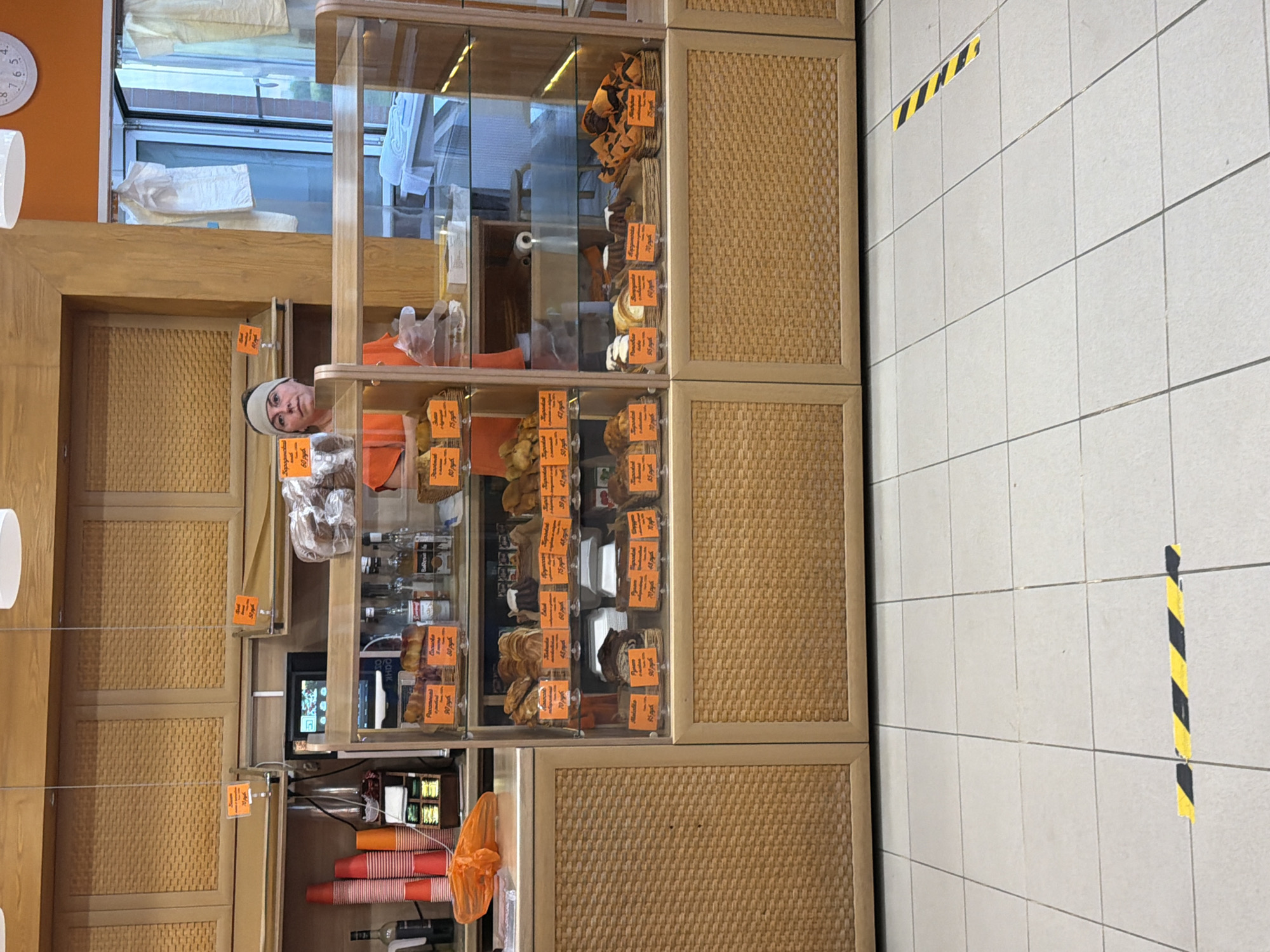 A woman wearing a grey headband and an orange shirt stands behind a glass display case filled with baked goods. The display case is in a bakery, with wooden cabinetry above and to the sides. The floor is tiled, with yellow and black hazard tape visible on the floor in the foreground. The woman appears to be a cashier or employee. The bakery is located in Shcherbinka, Russia.