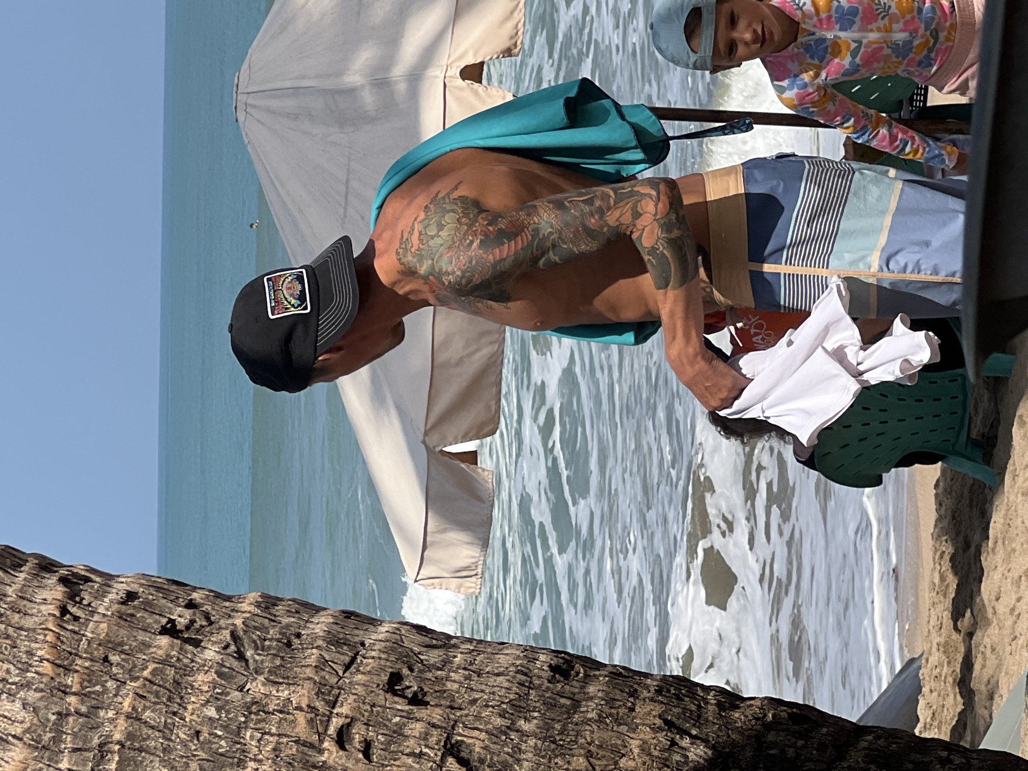 The image depicts a beach scene on a sunny day, possibly in Kuta, Indonesia. In the foreground on the left is the textured bark of a palm tree. To the right, a shirtless man with a large dragon tattoo on his left arm wears a black baseball cap and patterned swim shorts. A turquoise towel is draped over his right shoulder. He is holding a white t-shirt in his left hand. Behind him, a light-colored beach umbrella is partially visible. To the right of the man, a young child wearing a colorful rash guard and a blue baseball cap is looking to the left. The background features the ocean with gentle waves and a clear blue sky. The sand and the presence of beach umbrellas suggest a tourist or recreational area.