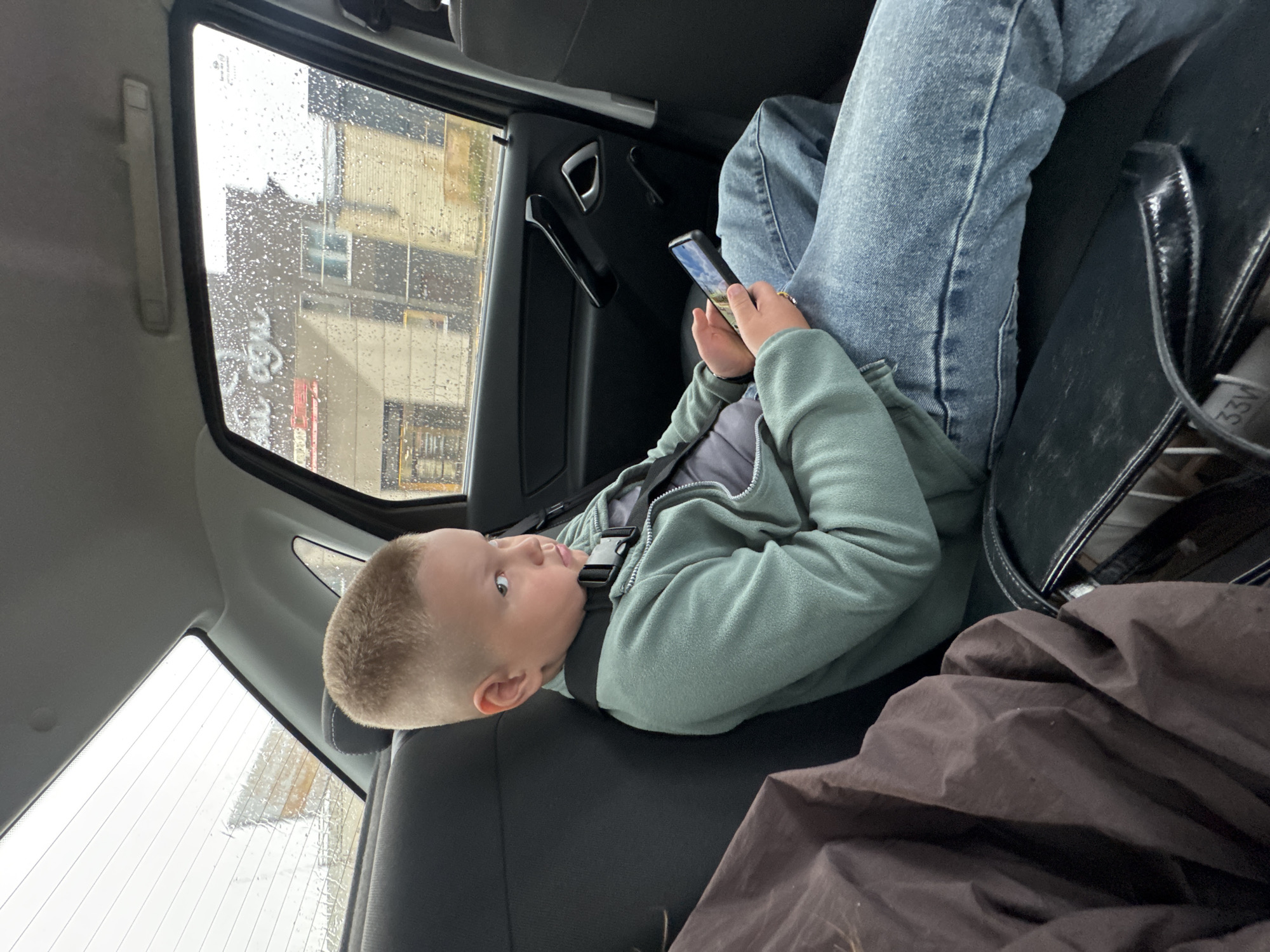 A young boy with blonde hair and a side part is seated in the back seat of a car, looking away from the camera with a contemplative expression. He is wearing a teal zip-up hoodie over a dark grey t-shirt and blue jeans. A black car seat strap is visible across his chest. The boy is holding a smartphone in his hands, its screen illuminated with an image. Outside the car window, a blurry cityscape is visible, with buildings and a sign that reads "Epic" in light-colored lettering. The window is speckled with raindrops, indicating overcast or rainy weather. A dark brown fabric bundle is placed on the seat next to the boy, and a black bag with a reflective strip and the number "33V" is visible in the foreground. The overall setting suggests a journey through the city of Chaikovsky, Russia, possibly on a damp day.