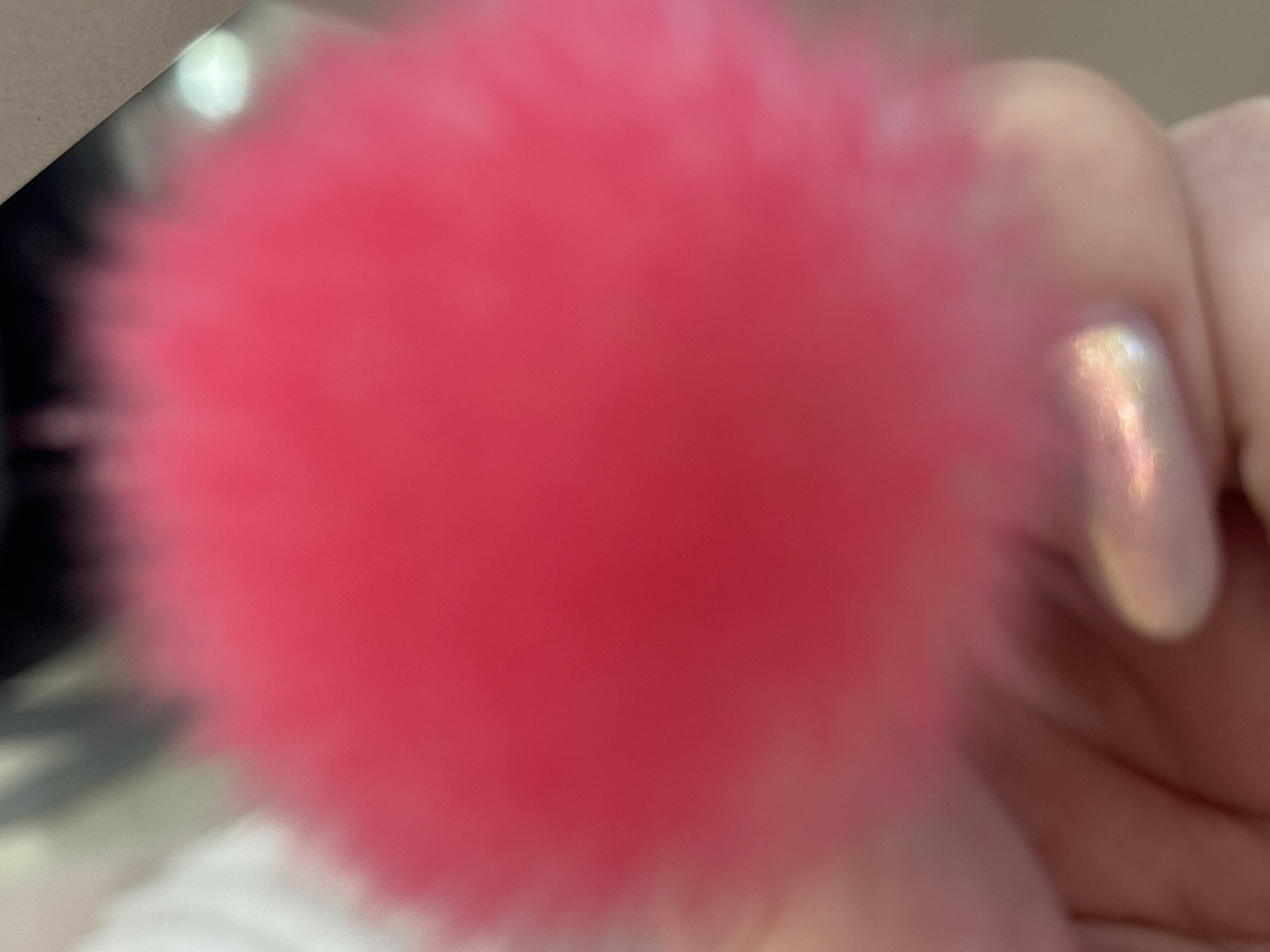 A close-up, out-of-focus photograph depicts a person holding a bright pink, fuzzy pom-pom. A hand with iridescent nail polish is visible to the right of the pom-pom. The background is blurry, showing white and dark grey tones. The location is not determinable from the image.