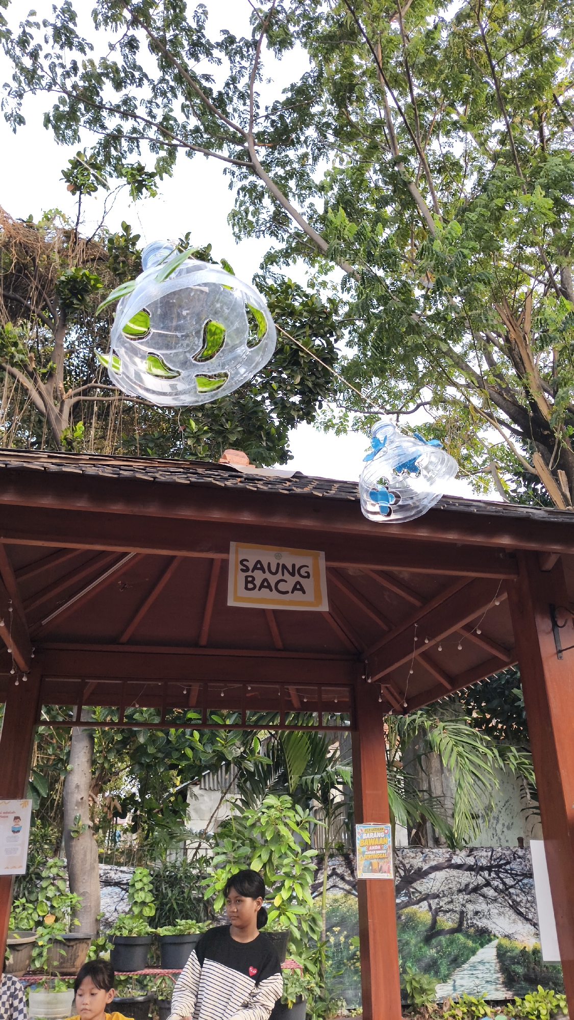 The image depicts an outdoor setting beneath trees, likely in South Jakarta, Indonesia. A wooden gazebo with a tiled roof is prominently featured, with a sign reading "SAUNG BACA" (Reading Corner) hanging from the roof. Two large, clear plastic bottles, repurposed as decorative elements, are suspended from the trees above the gazebo. These bottles appear to have been cut and shaped, with greenery placed inside one and blue accents in the other.
In the foreground, two young girls are seated. The girl on the left is wearing a yellow shirt and looking down. The girl on the right is wearing a black and white striped sweater and looking towards the right side of the frame. Behind them, various potted plants are visible, adding to the natural and somewhat rustic ambiance of the scene. The overall environment suggests a public space designed for leisure or community activities, possibly a park or a community garden.