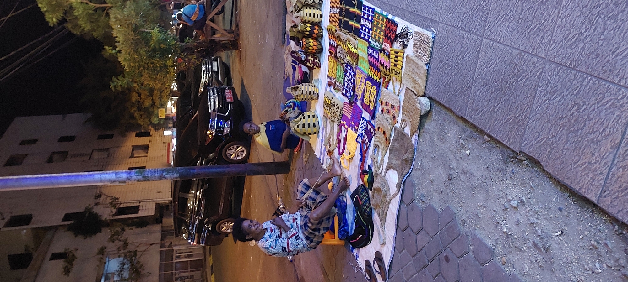 In Jayapura, Indonesia, two women are seen selling woven goods on a sidewalk at night. The woman on the left is wearing a patterned shirt and plaid shorts, sitting on an orange stool. The woman on the right is wearing a blue shirt with a yellow drape over her shoulders, sitting on the ground. They have laid out a variety of woven bags, placemats, and other items on a white cloth. Behind them, several cars are parked on the street, and a building is visible in the background. A street light with a blue glow illuminates the area. A third person, wearing a blue shirt and sitting on a wooden bench, is visible further in the background.