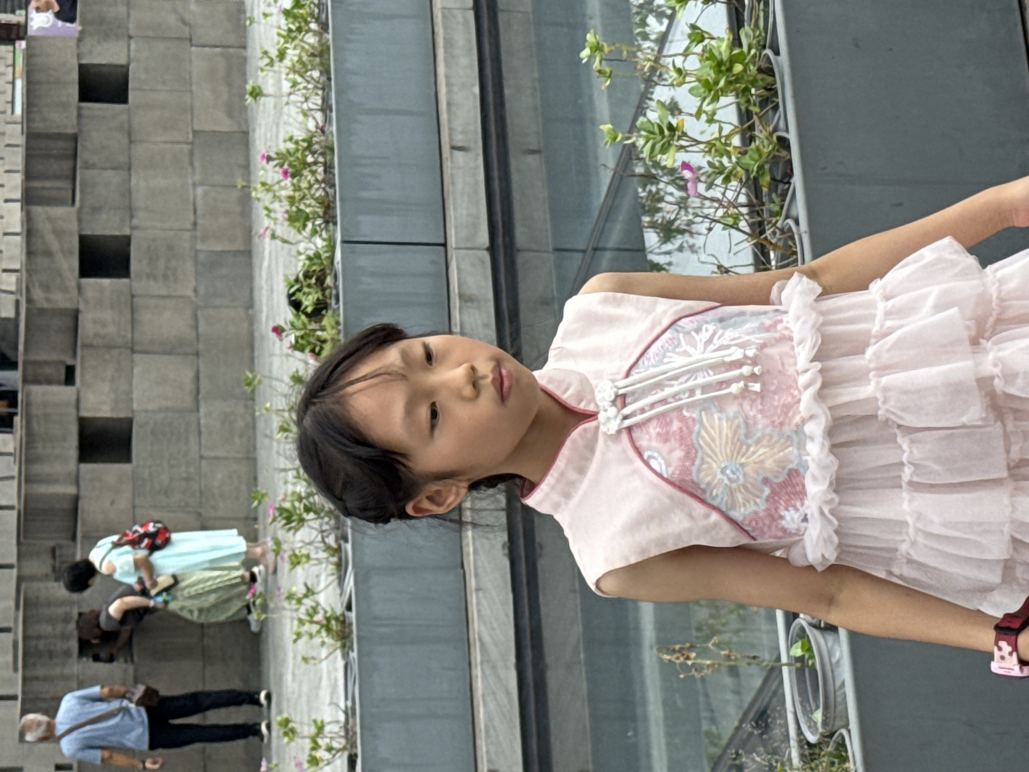 A young East Asian girl wearing a light pink sleeveless dress with ruffled layers and floral embroidery stands in the foreground. Her hair is pulled back in a braid, and she is looking down and to her right. She is wearing a pink digital watch on her left wrist. In the background, there are three people. A man in a blue t-shirt and dark pants stands on the left, facing away from the camera and looking at a wall. Two women are to his right. One woman, wearing a light green striped skirt and a dark t-shirt, is leaning in and looking at the wall. The other woman, wearing a light blue dress, stands behind her with a patterned backpack. They are all in front of a textured gray wall. The scene appears to be outdoors in a public space with planters of small green plants and pink flowers in the mid-ground and background.
