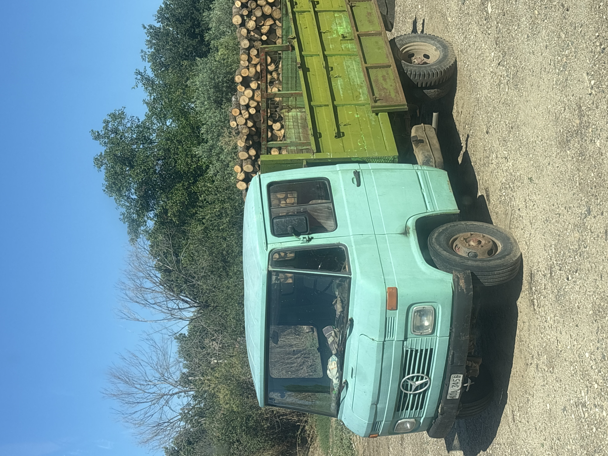 A turquoise Mercedes-Benz flatbed truck is parked on a gravel road in a rural area, possibly in or near Mikro Doukato, Greece. The truck is loaded with a large stack of cut logs in its green-painted bed. The truck's cab is also turquoise, and its windshield shows some items placed on the dashboard. The background consists of dense green foliage and trees under a clear blue sky. The overall scene suggests a logging or forestry operation in a sunny, outdoor environment.