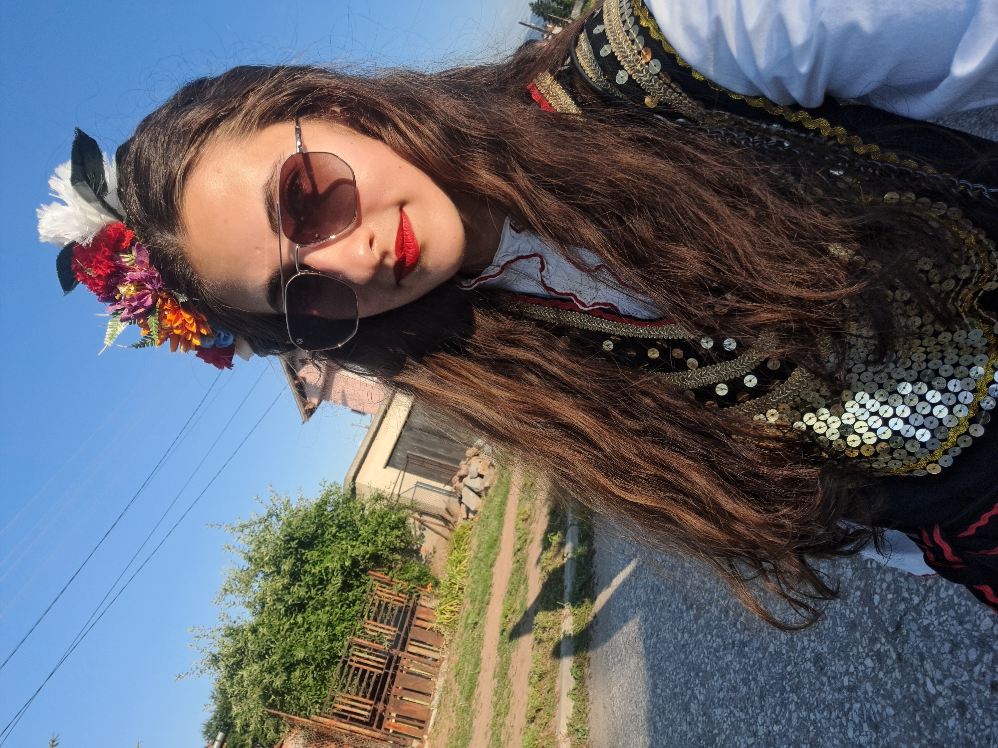 A young woman with long brown hair adorned with a flower crown and wearing a traditional, sequined vest over a white shirt and red and black patterned skirt poses for a selfie outdoors. She is wearing aviator sunglasses and bright red lipstick. The background shows a dirt path, a fence, trees, and buildings under a clear blue sky. The lighting suggests it is daytime, likely late afternoon due to the long shadows. The location is an outdoor, rural or suburban setting, possibly a village.