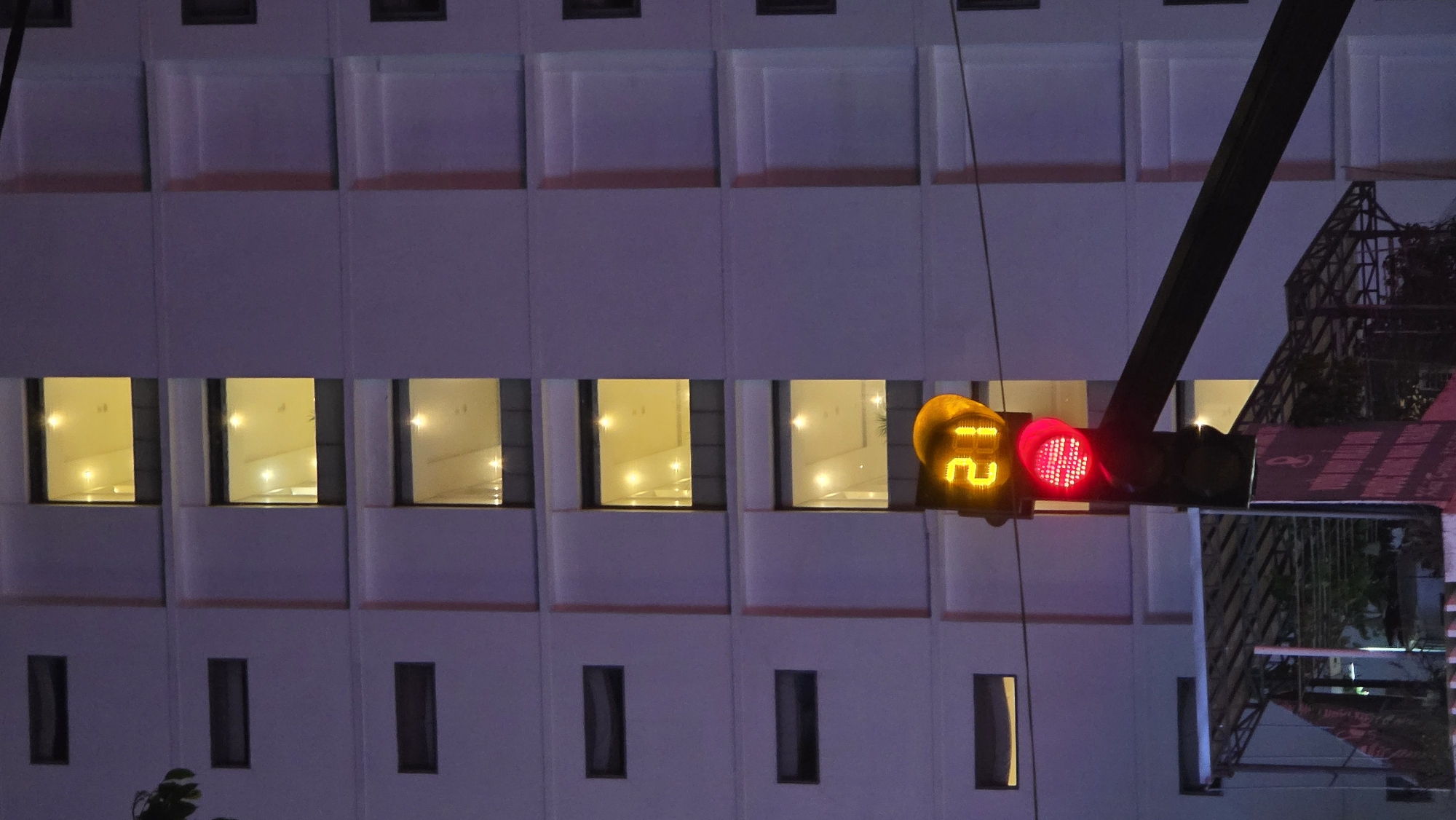 The image depicts a traffic light in Nha Trang, Vietnam, showing a red light with a countdown timer displaying "21". The traffic light is positioned in front of a building facade featuring a grid of windows. Some windows are illuminated from within, suggesting occupied rooms. The building is likely located in an urban area, as evidenced by the presence of street infrastructure. The photograph was taken at night, as the lighting is artificial and the sky is dark. A metallic structure, possibly part of a nearby construction or decorative element, is visible behind the traffic light.