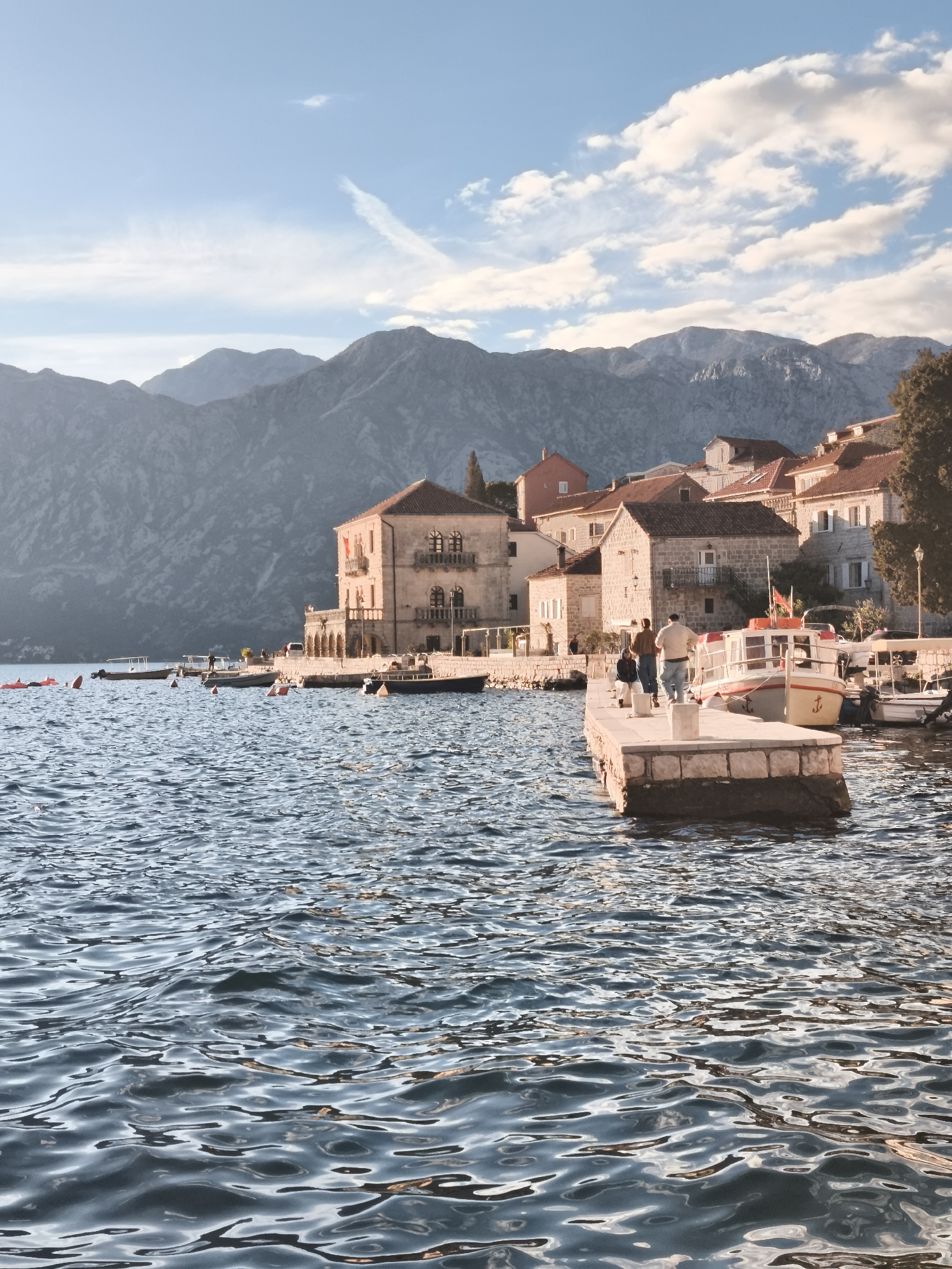 The image shows a waterfront scene in a coastal town. In the foreground is rippled water, reflecting sunlight. A stone pier extends into the water, with three people walking on it; an adult male and female, and a child. Several boats are docked along the pier. Behind the pier, the town's buildings are visible, with terracotta-tiled roofs and stone or stucco facades. The buildings vary in height and style, suggesting a historical settlement. In the background, large, grey mountains rise dramatically, contrasting with the blue sky dotted with clouds. The overall lighting suggests it is either morning or late afternoon.